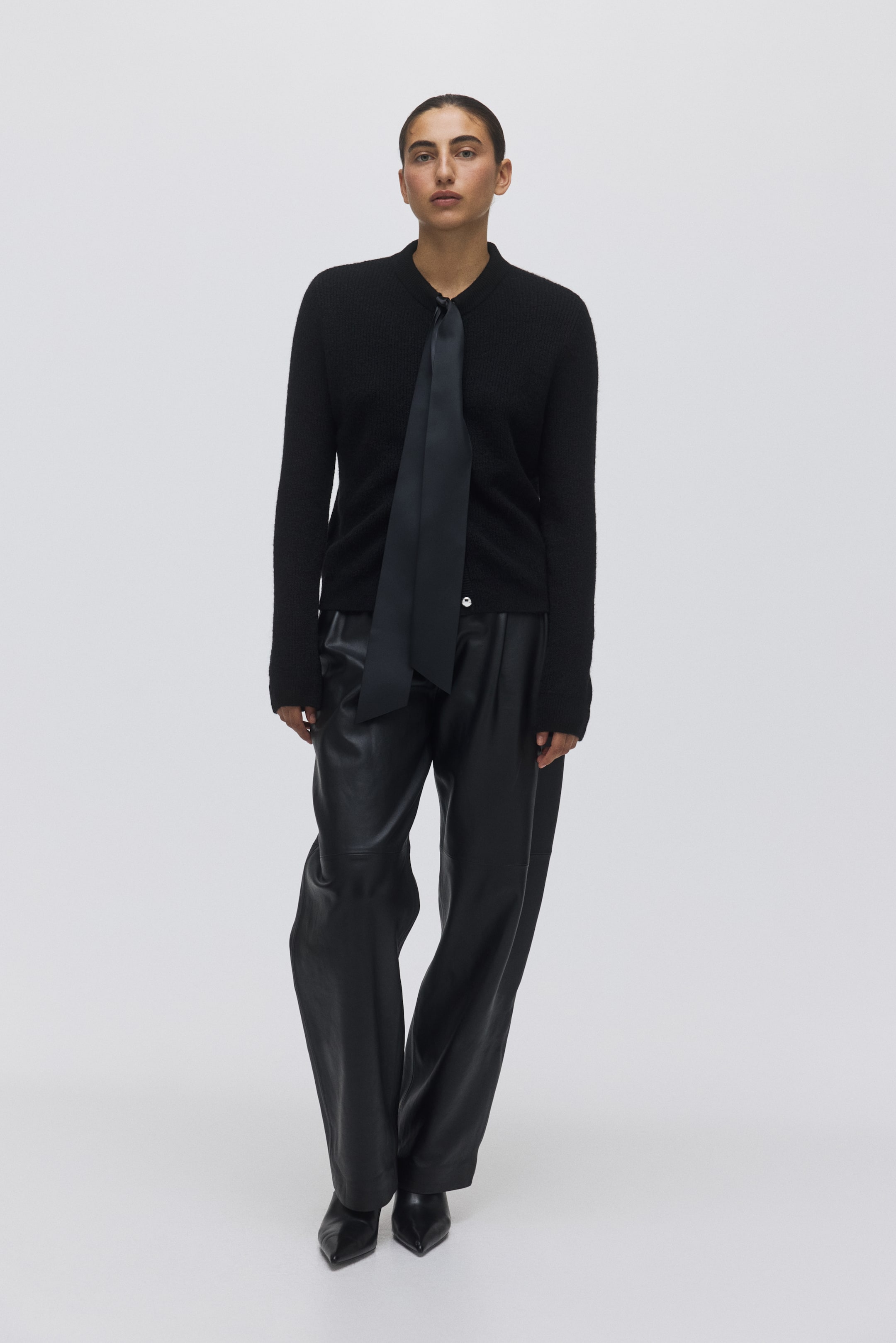 Büyük resmi görüntüle: Bir siyah örgü hırka sunuluyor, yaka kısmı yok ve boynun etrafına gevşek bir şekilde bağlanmış, hırkanın ön kısmına sarkan düzgün siyah bir kuşakla öne çıkıyor. Siyah pileli deri pantolonlar ve siyah sivri burunlu ayakkabılarla eşleştirilmiş, tüm kombin öne doğru bakıyor.