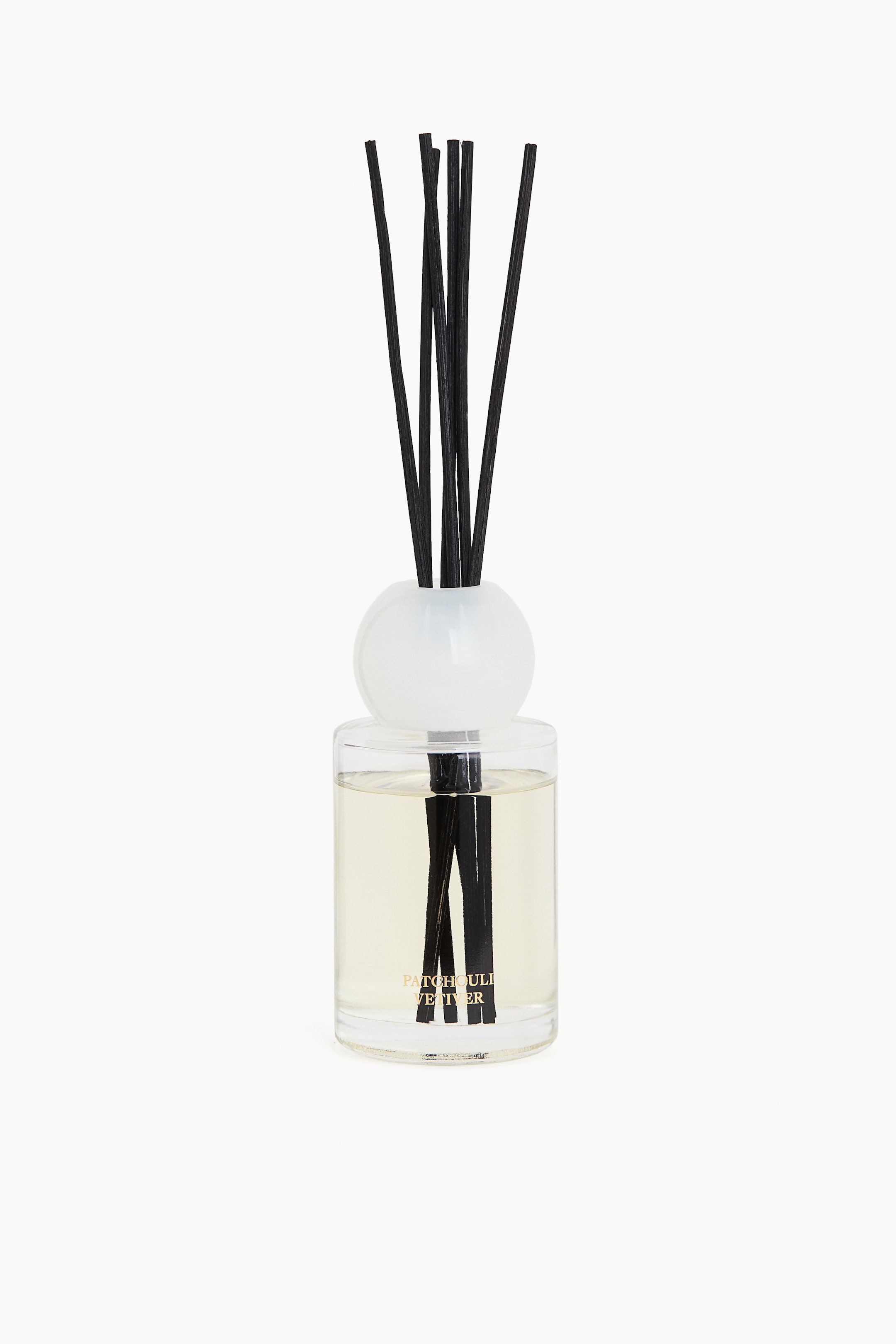 View larger image: A reed diffuser is placed upright, facing forward. It features a clear cylindrical glass bottle containing a light yellow liquid, with 'PATCHOULI VETIVER' printed in gold on the front, and topped with a spherical white glass cap. Several black reeds emerge from the cap and are immersed in the liquid.