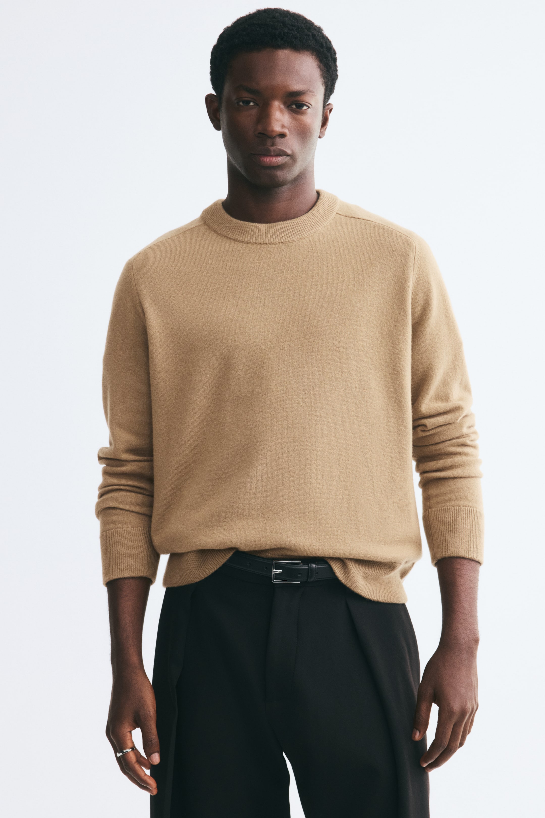 Agrandir l'image: Un pull beige à col rond est porté par le mannequin, avec un col, des poignets et un ourlet côtelés. Le pull est stylisé avec l’ourlet avant rentré de manière décontractée dans un pantalon noir maintenu par une ceinture noire.