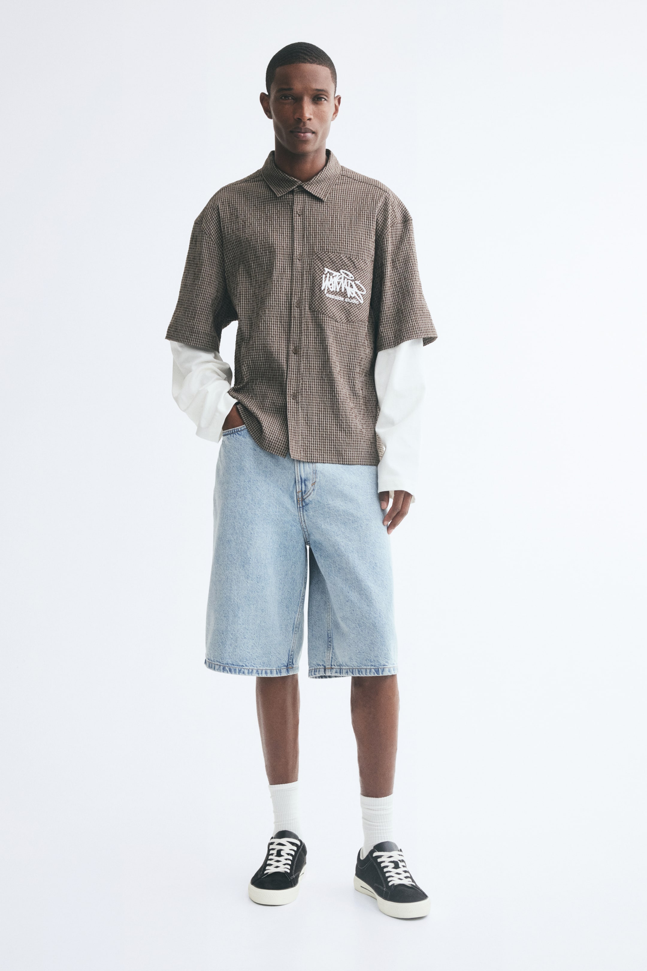 Vezi imaginea mai mare: Șorturi din denim bleu deschis, cu croială lejeră și largă, purtate de model, care își sprijină o mână în buzunarul din față. Șorturile până la genunchi sunt asortate cu o cămașă maro cu imprimeu, cu mânecă scurtă și nasturi, purtată peste o cămașă albă cu mânecă lungă, șosete albe și sneakers negri.