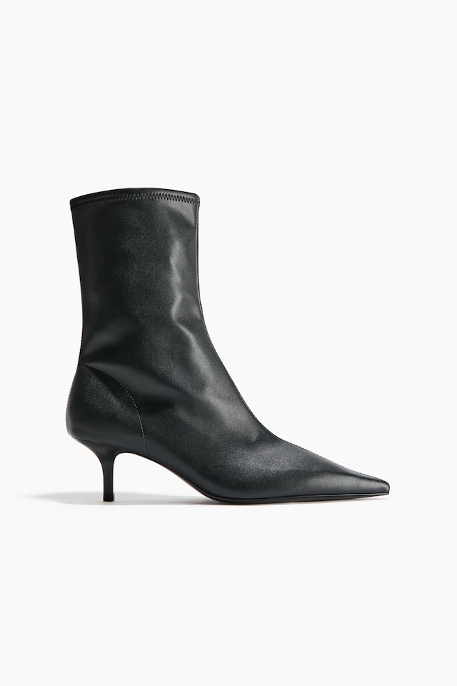 A black leather ankle boot is facing right in a side profile, featuring a highly pointed toe with a central seam and a low kitten heel. The boot is displaying a fitted, sock-style shaft that extends above the ankle with a clean-finished top edge. - 1