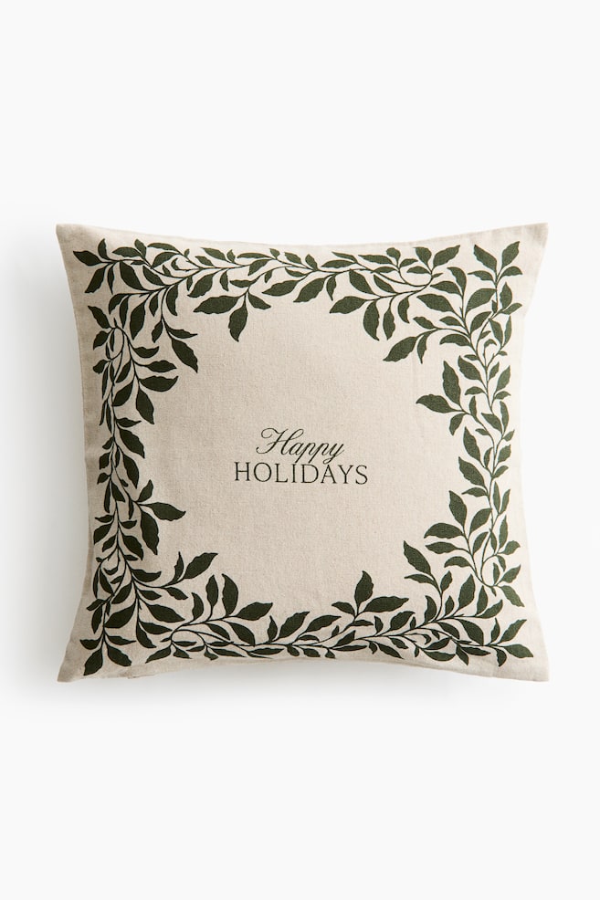 A square cushion cover in a beige linen-blend fabric is shown from the front, featuring a dark green leafy vine border printed along the edges. The centre displays the words "Happy HOLIDAYS" printed in a dark green cursive and serif font. - 1
