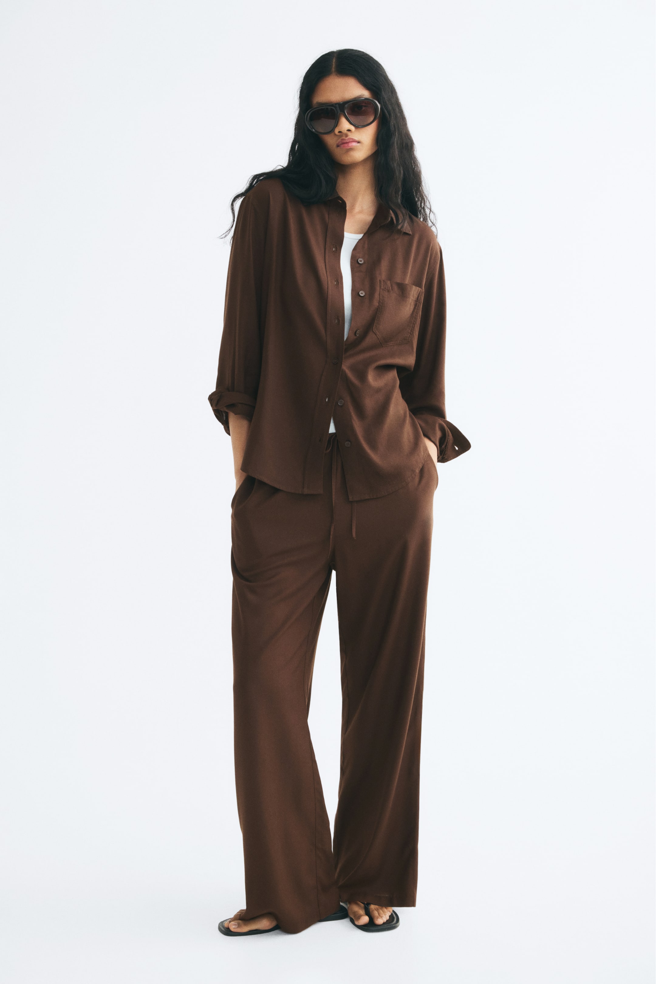 View larger image: A brown, long-sleeved linen-blend shirt featuring a collar, front button closure, and a single patch chest pocket is worn open by the model. The shirt is layered over a white top and paired with matching brown wide-leg trousers.