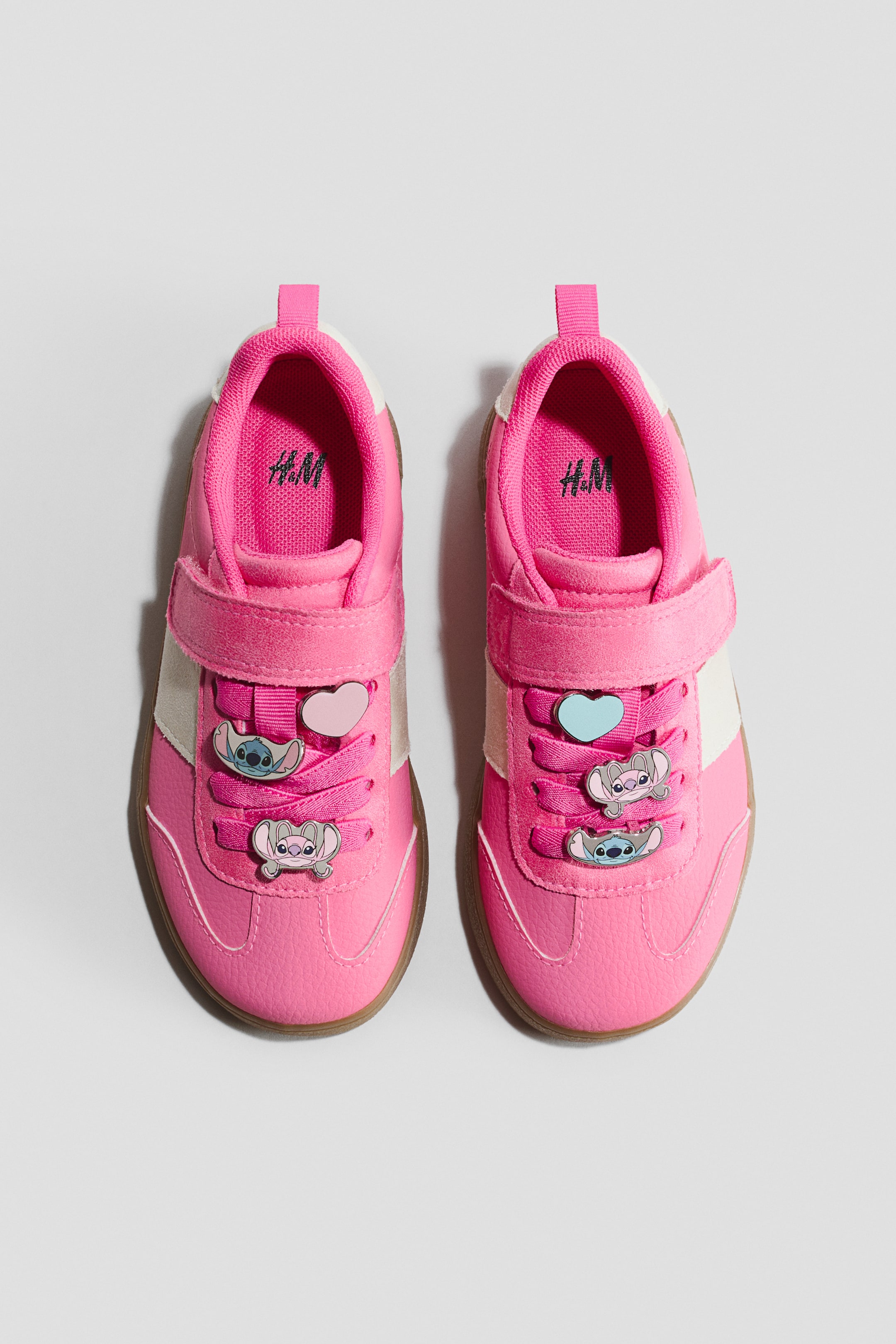 View larger image: Motif-detail sneakers are shown from the front, showcasing a vibrant pink color with white side stripes and brown soles. The sneakers feature pink laces adorned with character and heart-shaped charms, secured by a pink hook-and-loop strap.