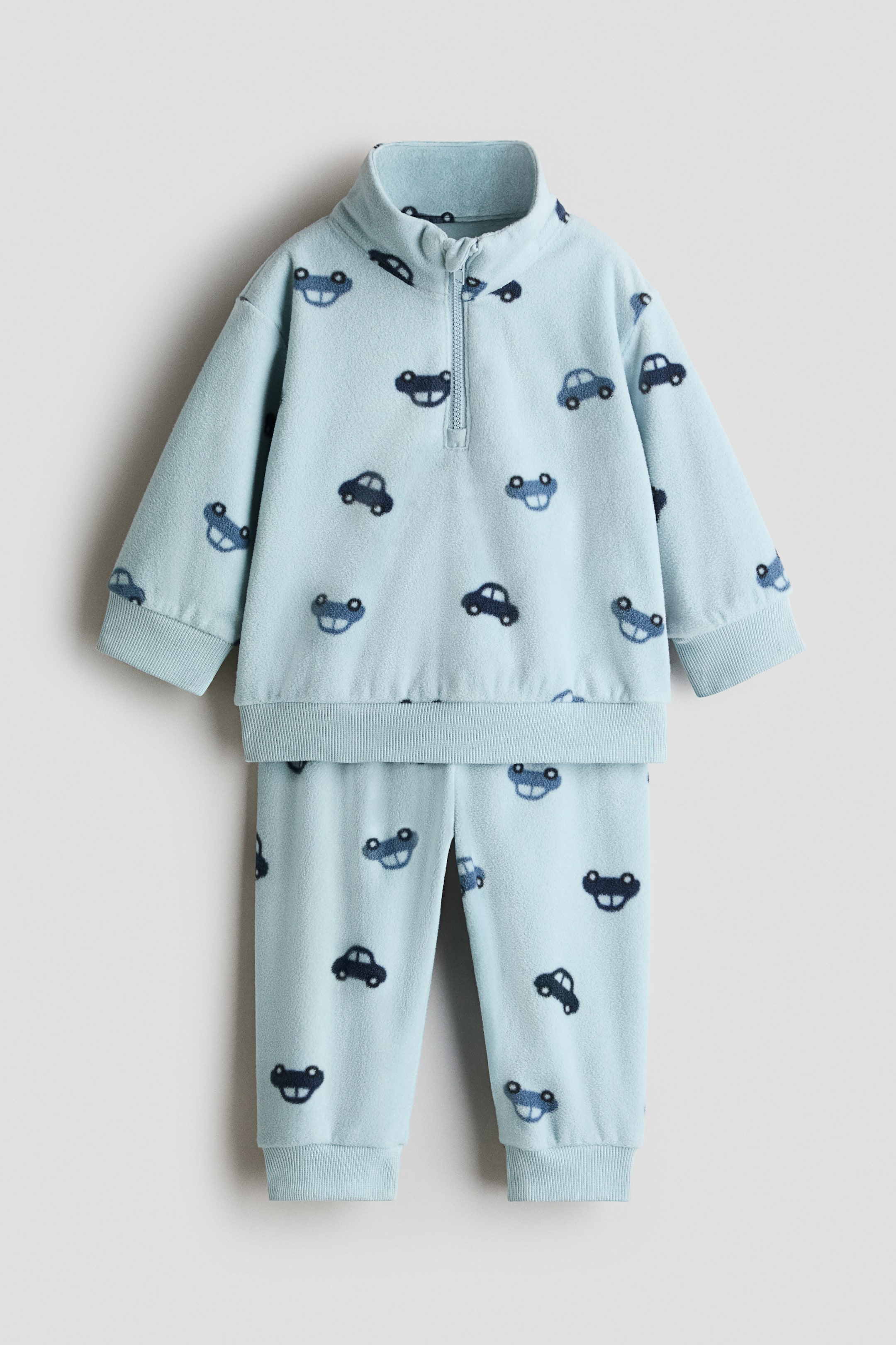 View larger image: A light blue fleece two-piece set is shown from the front, featuring an all-over pattern of small blue cars. The sweater is designed with a stand-up collar, a half-zip closure, dropped shoulders, and ribbed cuffs and hem, paired with matching pants that have ribbed ankle cuffs.