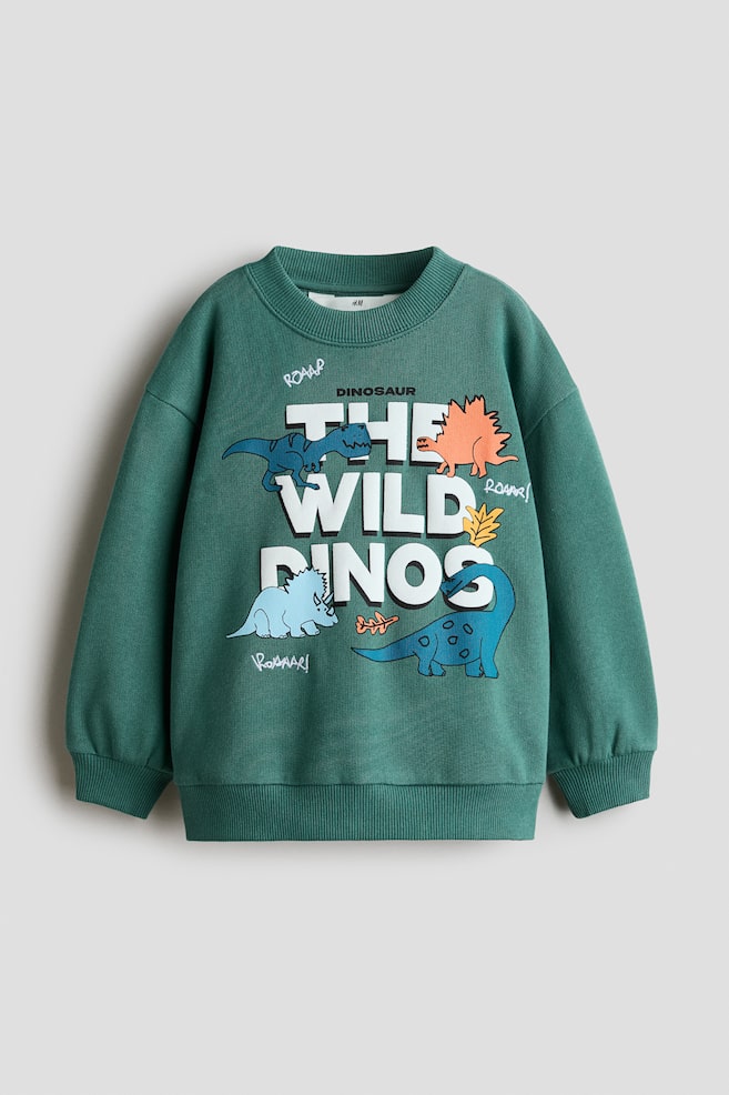 A green sweatshirt is shown from the front, featuring a round ribbed neckline, dropped shoulders, and long sleeves with ribbed cuffs and a ribbed hem. The front displays a printed motif with the text "THE WILD DINOS" in bold white block letters, surrounded by colourful cartoon dinosaur illustrations. - 1