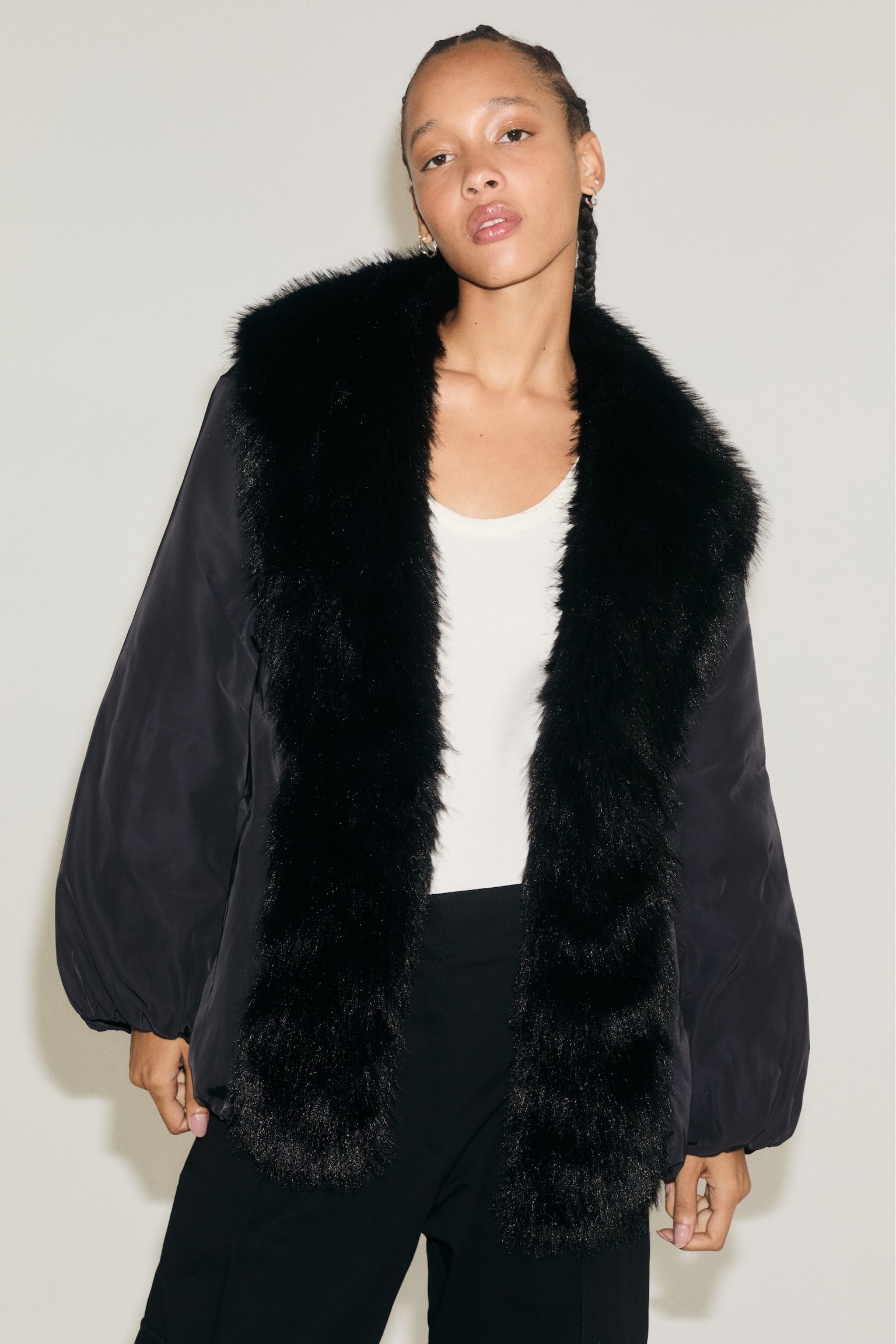 A black padded jacket is shown from the front, featuring a voluminous black faux-fur collar that extends down the front opening. The jacket is cinched at the waist with a matching self-tie fabric belt and showcases long sleeves with gathered cuffs and a puffed hem. - 1