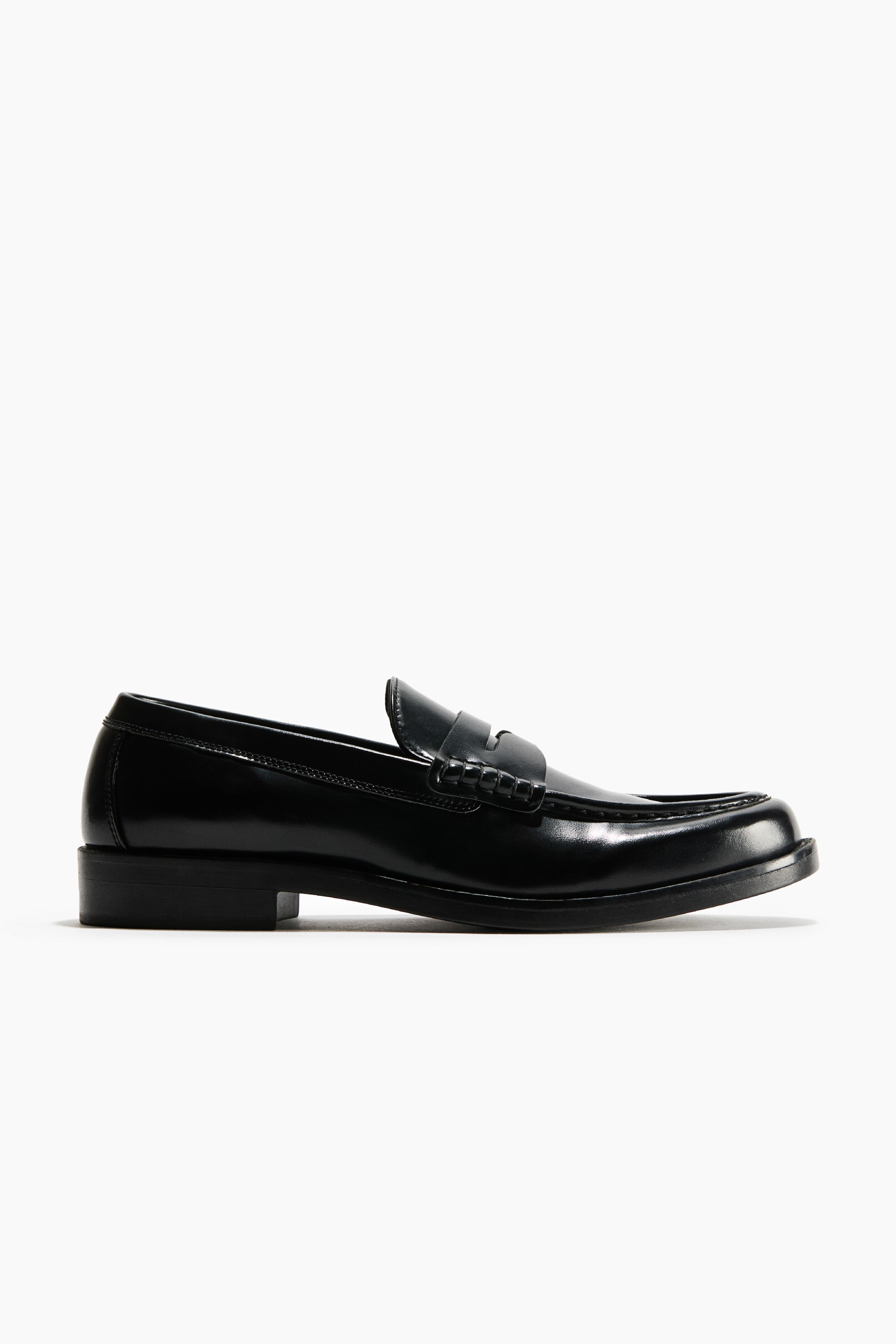 View larger image: A black leather penny loafer is resting in a side-profile view facing right, showcasing a polished finish and a classic leather saddle strap across the vamp. It features moc-toe stitching and a low block heel with a matching black sole.