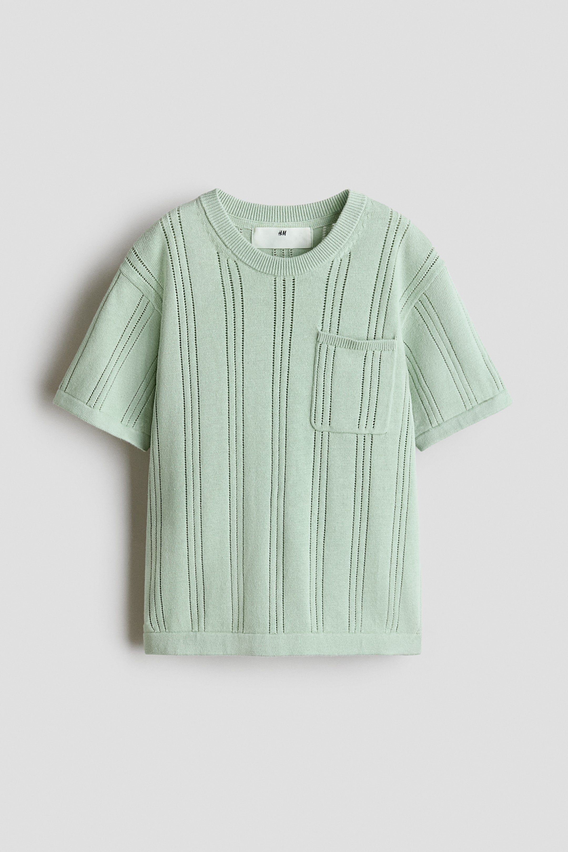 Größeres Bild anzeigen: Ein hellgrünes, strukturiert gestricktes T‑Shirt wird von vorne gezeigt. Es hat einen runden, mit Rippenbündchen eingefassten Ausschnitt sowie leicht überschnittene Kurzärmel. Vertikale Ajourlinien zieren das gesamte Kleidungsstück, und eine aufgesetzte Brusttasche befindet sich auf der linken Seite.