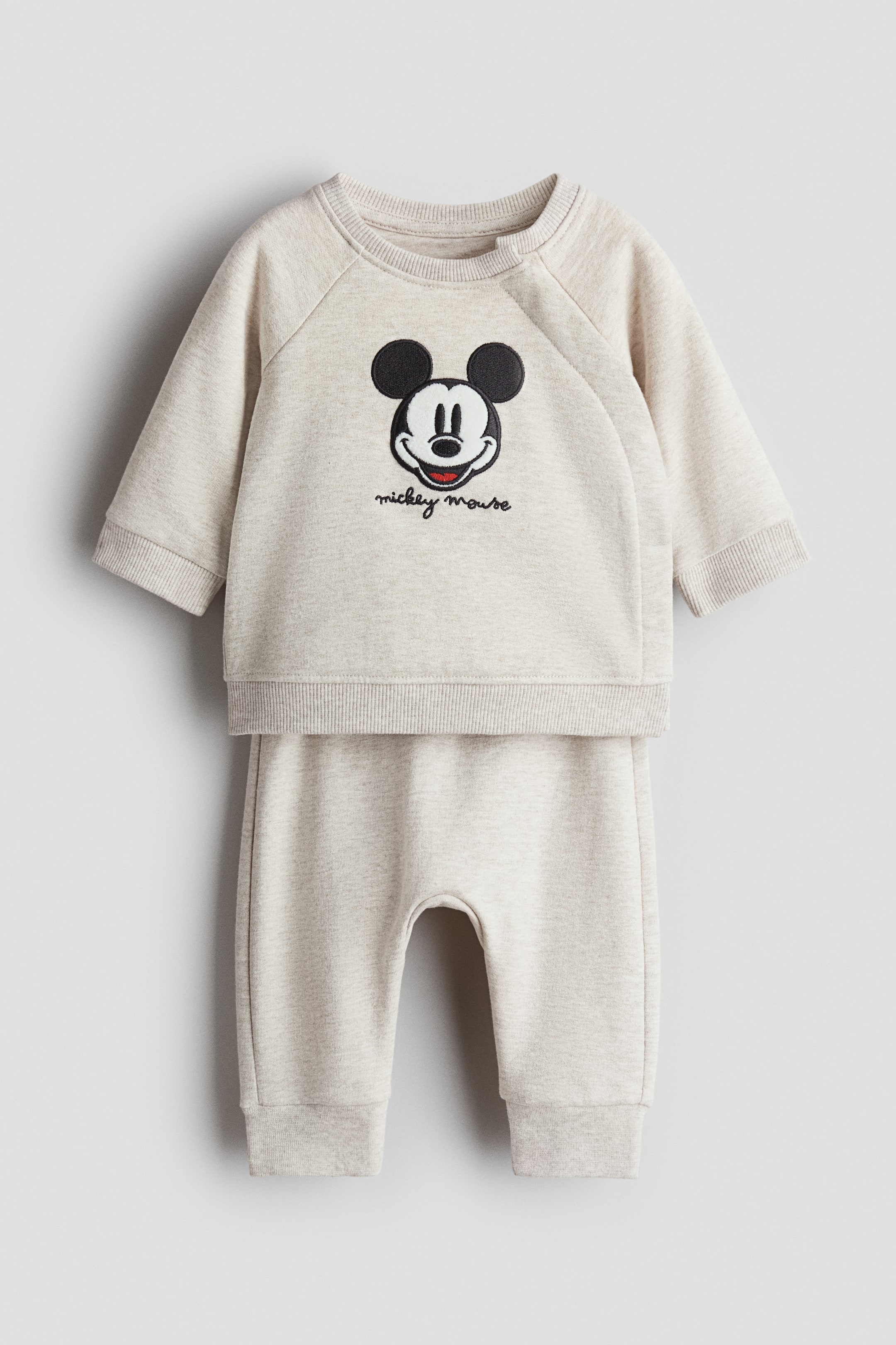 Agrandir l'image: Un ensemble bébé deux pièces en gris clair chiné est présenté de face. Le sweat-shirt à col rond arbore une tête de Mickey Mouse brodée et le texte "mickey mouse" sur le devant, un col côtelé avec fermeture à pression, des manches raglan avec poignets côtelés, et un ourlet côtelé. Le pantalon de jogging assorti présente des poignets côtelés et une entrejambe basse visible.