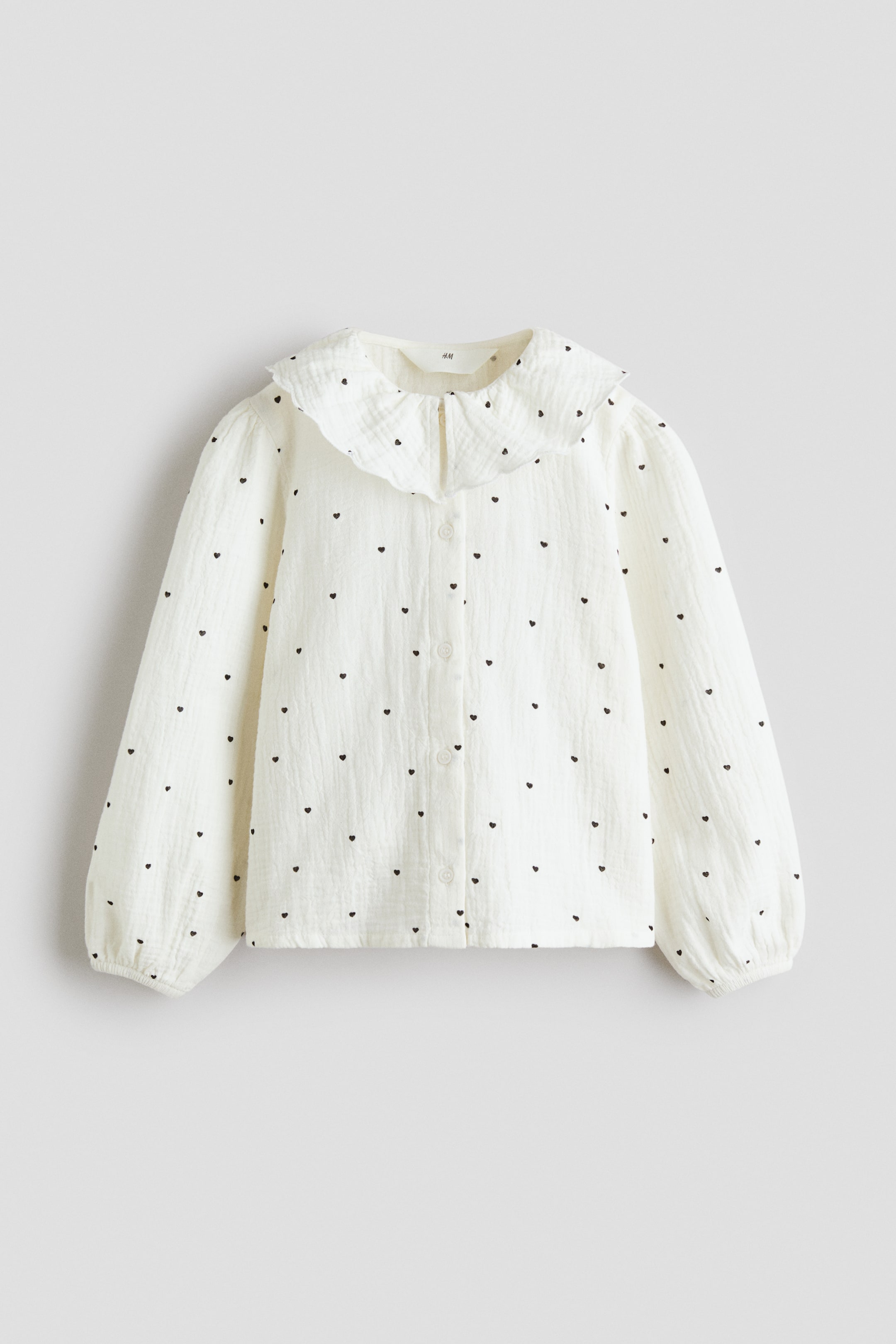 View larger image: An off-white blouse is showcasing a frontal view, highlighted by a ruffled collar and a complete button-front closure. It is crafted with long puffed sleeves, gathered cuffs, and features an all-over pattern of small black hearts on a textured fabric.