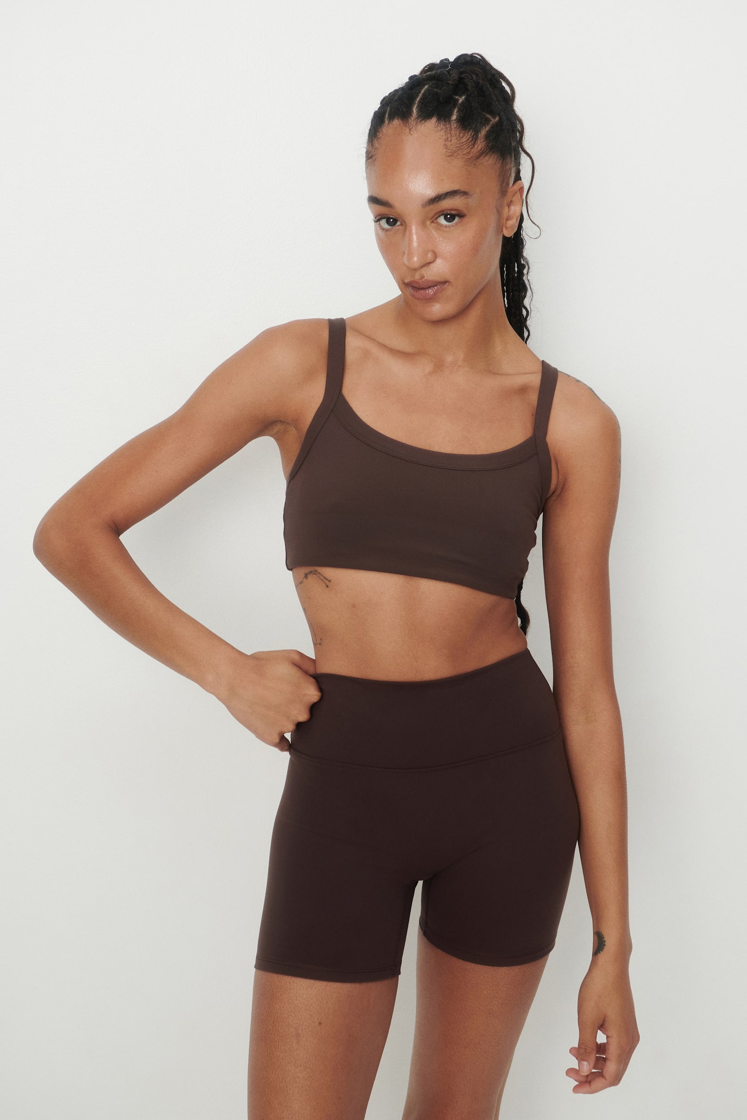 A brown light-support sports bra with wide straps and a scoop neckline is being worn by the model, who is also wearing matching brown cycling shorts and resting her left hand on the waistband. - 1