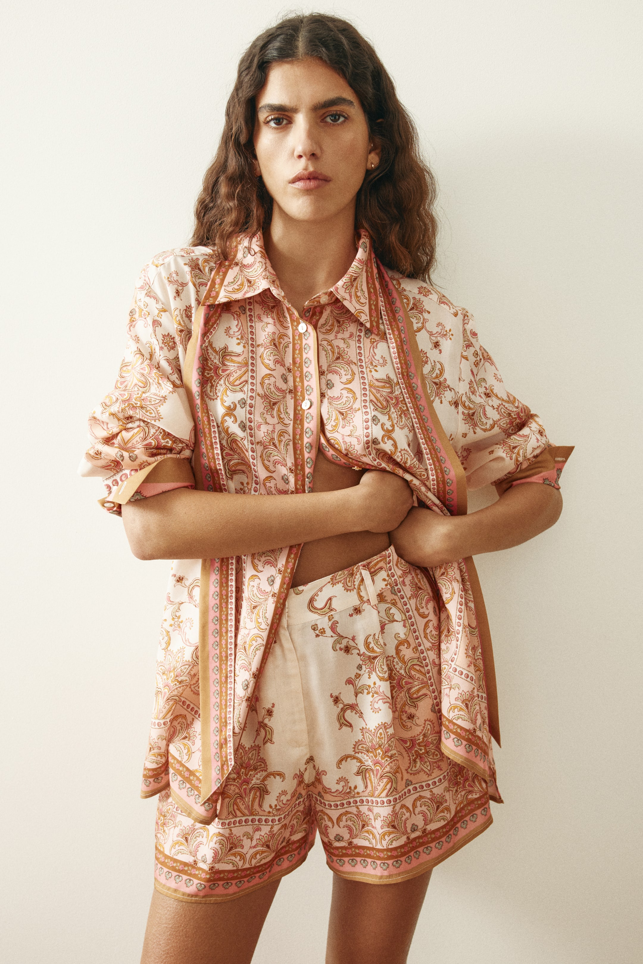 Visualizza immagine più grande: Il set coordinato di camicia e shorts in tonalità chiare è presentato frontalmente, mostrando un motivo paisley e a righe dettagliato nelle sfumature di rosa, arancione e marrone. La camicia con colletto presenta maniche lunghe arrotolate ai polsini e un'apertura frontale, mentre gli shorts coordinati a vita alta completano l'ensemble dall'aria rilassata.