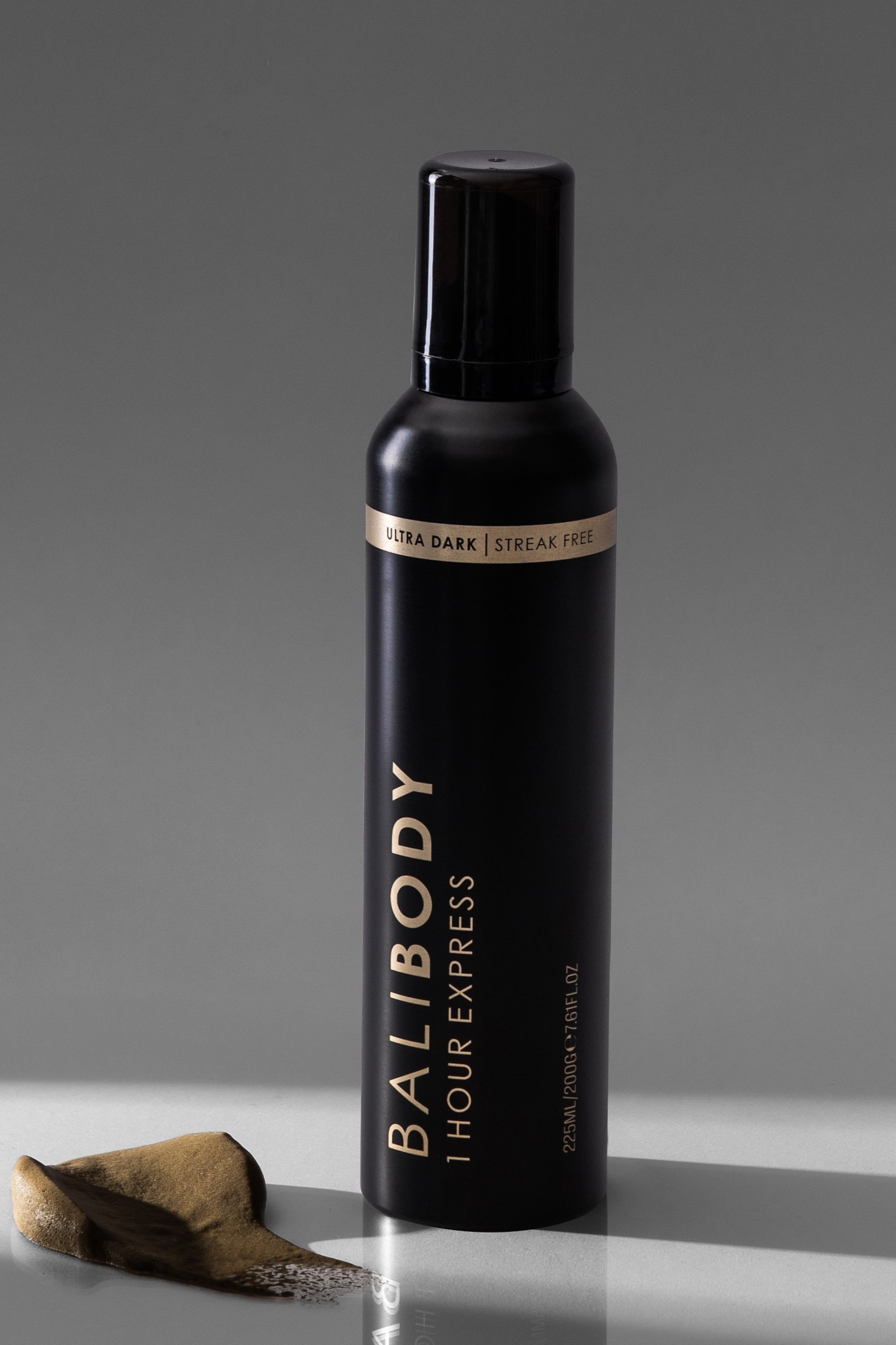 Visa större bild: En matt svart cylindrisk BALIBODY-flaska med en glänsande svart kork står upprätt, med etiketten framåt och något till vänster. Guldbokstäver stavar "BALIBODY 1 HOUR EXPRESS" och "225ML/200G/7.61FL.OZ" längs sidan, medan ett guldband indikerar "ULTRA DARK | STREAK FREE" nära toppen. En mörkbrun, skummig substans samlas på ytan bredvid flaskans bas.