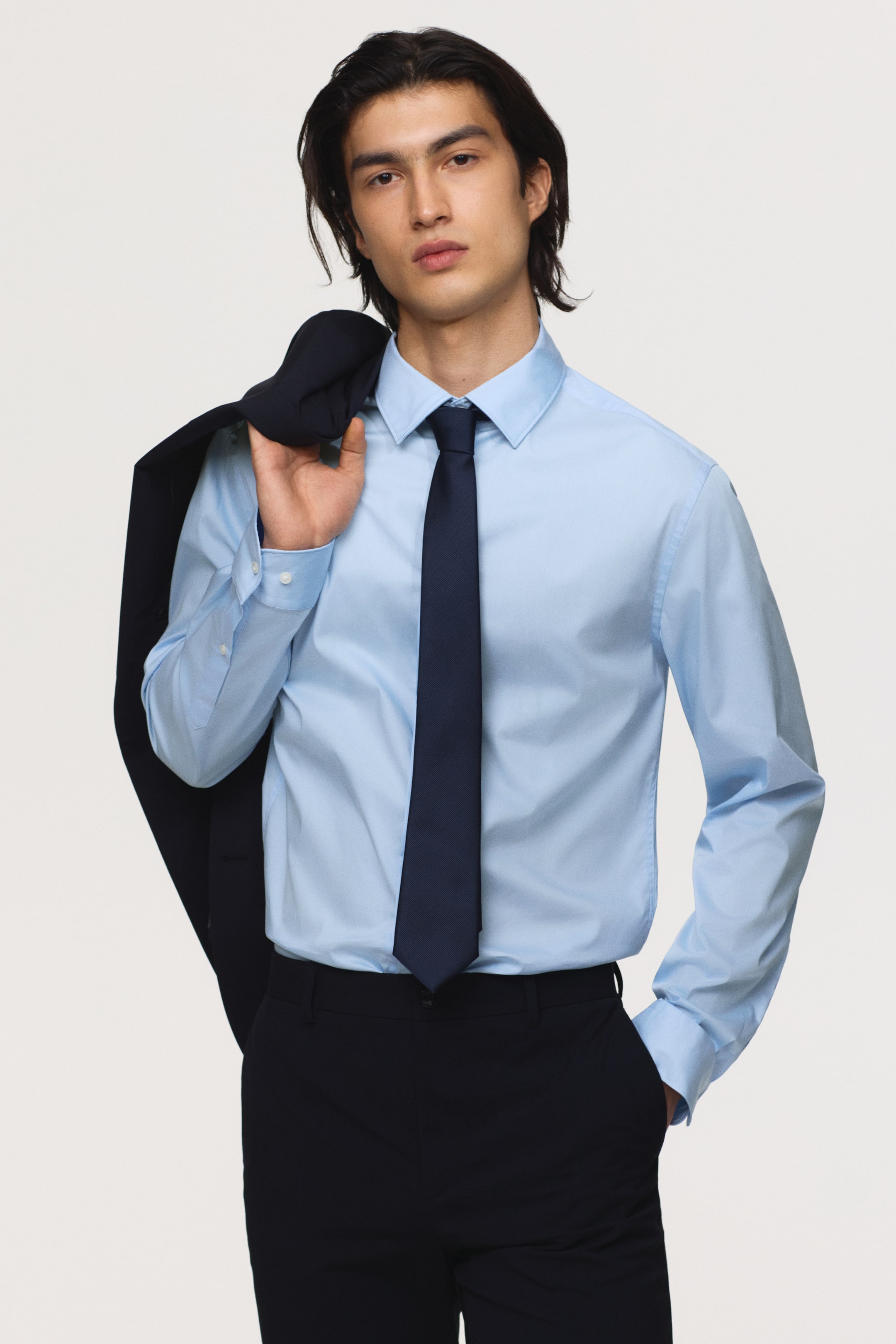 View larger image: A light blue long-sleeved dress shirt, showcasing a classic collar and buttoned cuffs, is presented from a slightly angled front view. It is styled with a solid navy tie and dark trousers, while a dark jacket is draped over the left shoulder.