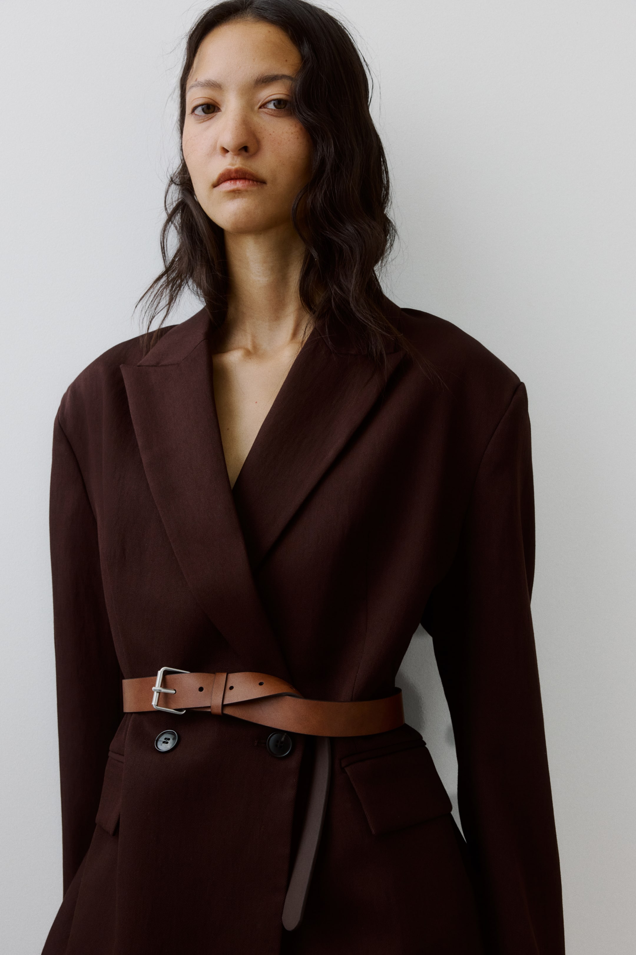 Agrandir l'image: Un blazer croisé marron foncé est doté de larges revers en pointe et de deux boutons sombres à l'avant, avec une poche à rabat visible sur le côté droit. Une ceinture en cuir marron avec une boucle argentée resserre le blazer à la taille.