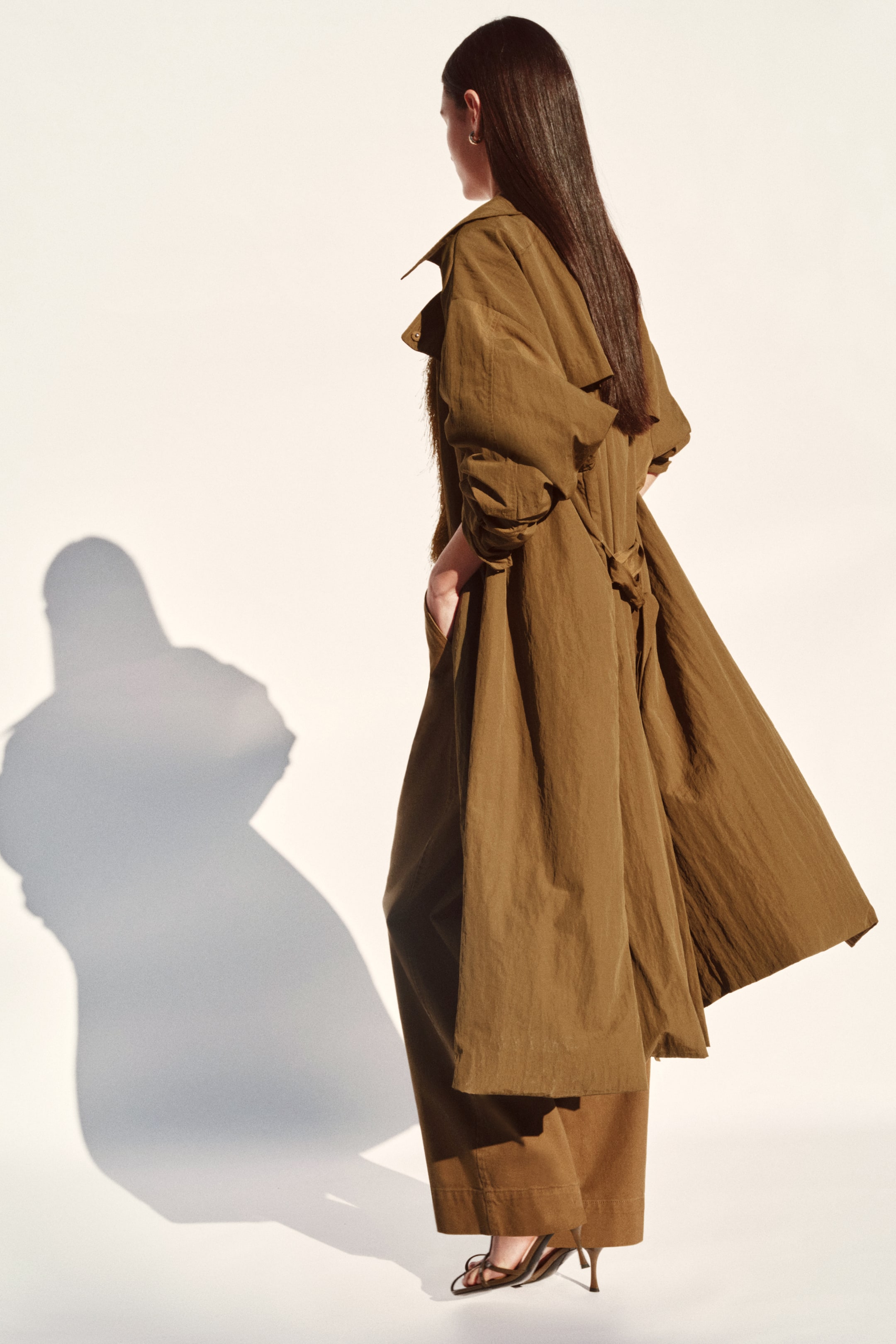 View larger image: An olive green long coat showcases a flowing, ankle-length silhouette, viewed from the back and side. It highlights a crinkled texture, a spread collar, raglan sleeves, a fabric belt tied at the back, and side pockets, layered over matching wide-leg trousers.