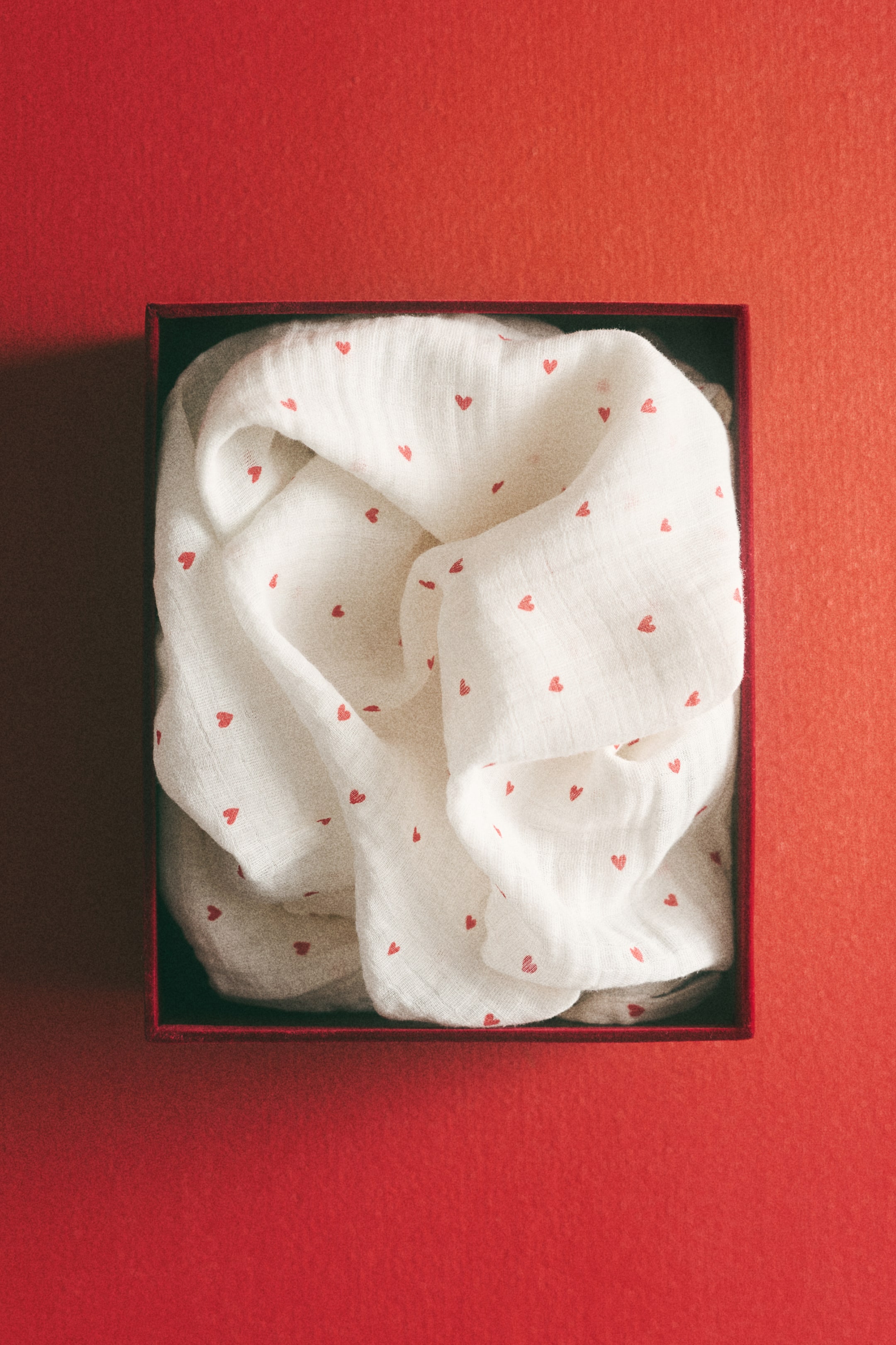 View larger image: A white crinkled fabric, adorned with small red hearts, is gently folded inside a red rectangular box.