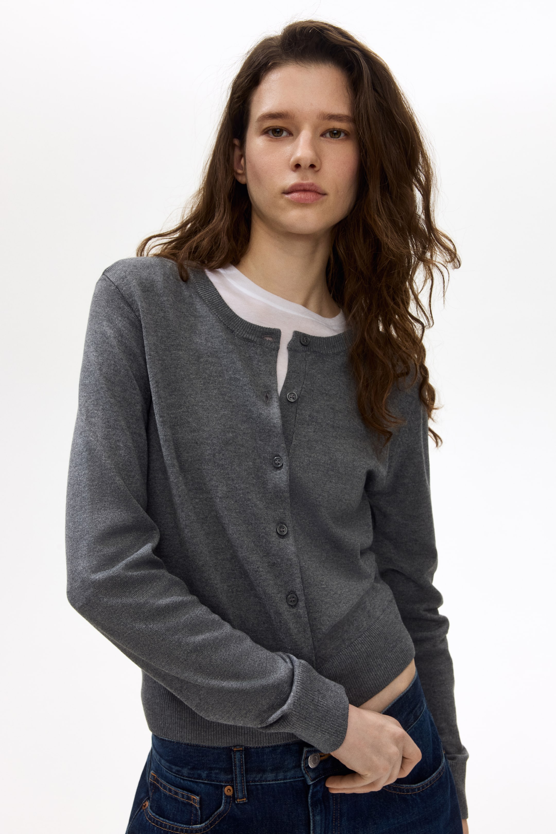 View larger image: Presenting a grey cardigan from the front, it features a round neckline and a button-front closure with dark grey buttons. The cardigan is styled open over a white t-shirt, highlighting its long sleeves with ribbed cuffs and a ribbed hem.
