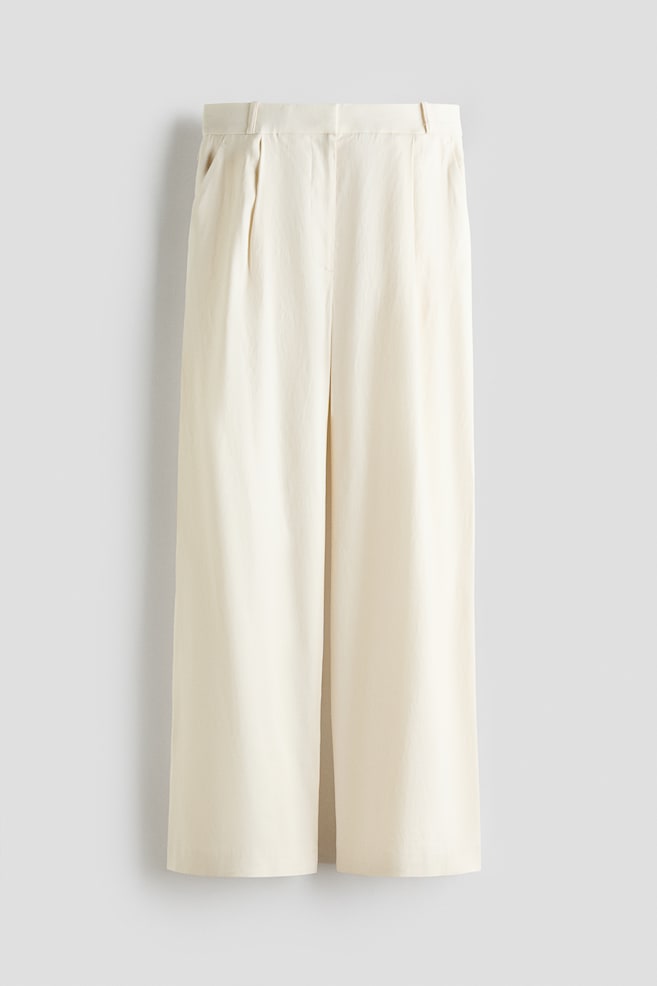 Pantalon crème à jambes larges présenté de face, avec une ceinture plate dotée de passants pour ceinture. Deux plis marqués façonnent chaque côté de l'avant, avec des poches latérales visibles, et les jambes tombent dans une coupe large et droite. - 1