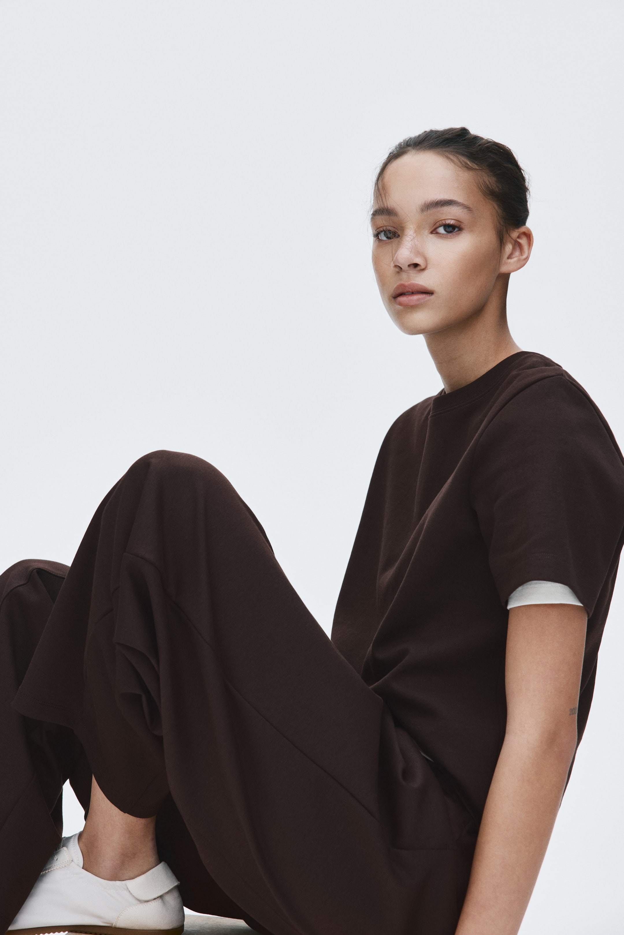 View larger image: A dark brown two-piece set is being presented, comprising a short-sleeved, relaxed-fit crewneck sweatshirt and wide-leg trousers. A white undershirt is visibly layering beneath the sweatshirt's sleeves. The loose-fitting trousers are showing a flowing design, gathering softly at the legs as the model is sitting, emphasizing their drape.