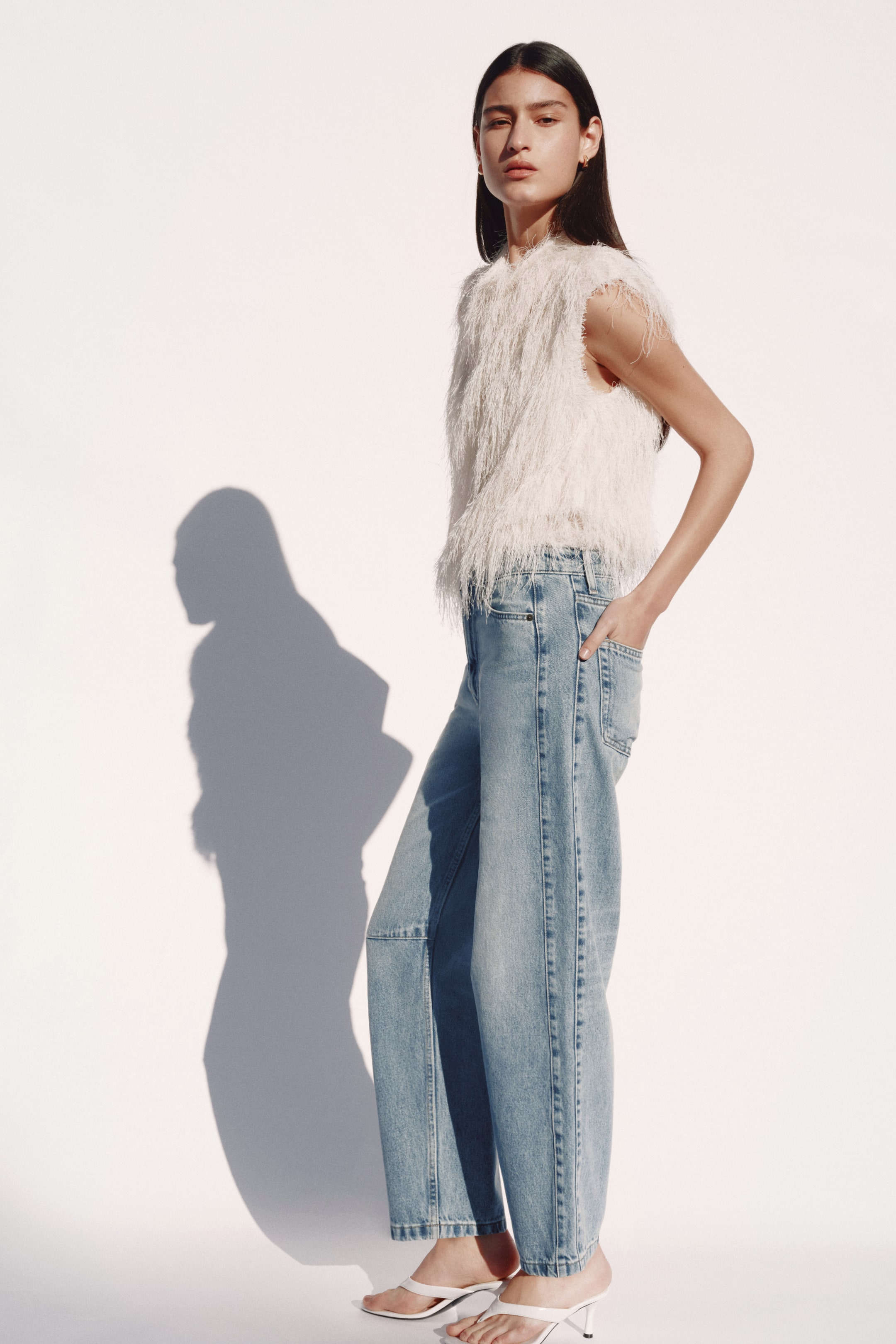 View larger image: Light blue 5-pocket jeans with a high waist and zip fly are worn by the model. The jeans are characterised by a voluminous, curved leg with knee darts and forward-facing side seams, tapering to an ankle length, and the model places her left hand into a front pocket.