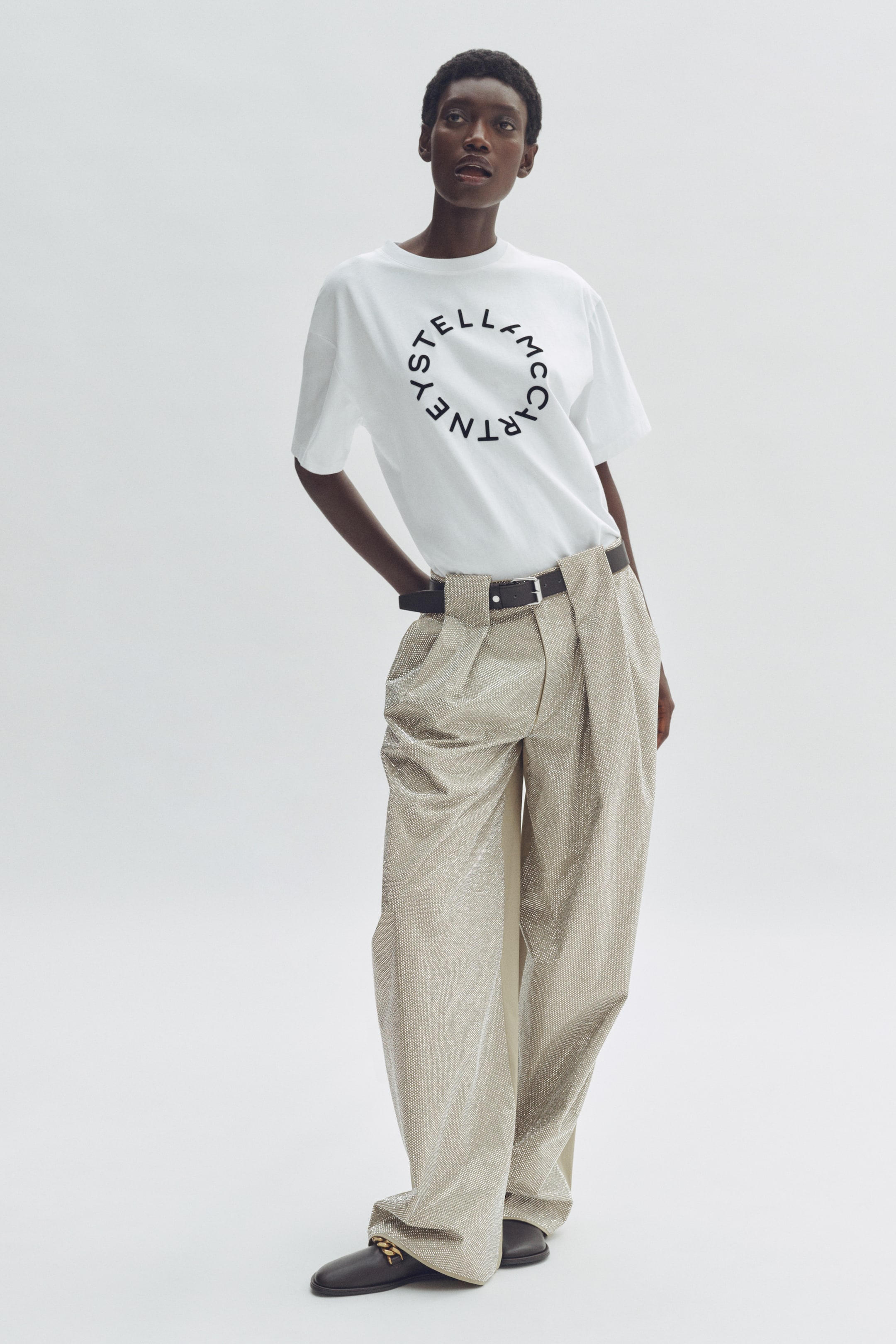 View larger image: A white short-sleeved t-shirt, featuring a black circular 'Stella McCartney' logo on the chest, is paired with high-waisted, wide-leg shimmering light gold trousers with front pleats. A black belt with a silver buckle cinches the trousers, while black loafers adorned with a gold-toned chain detail complete the ensemble.