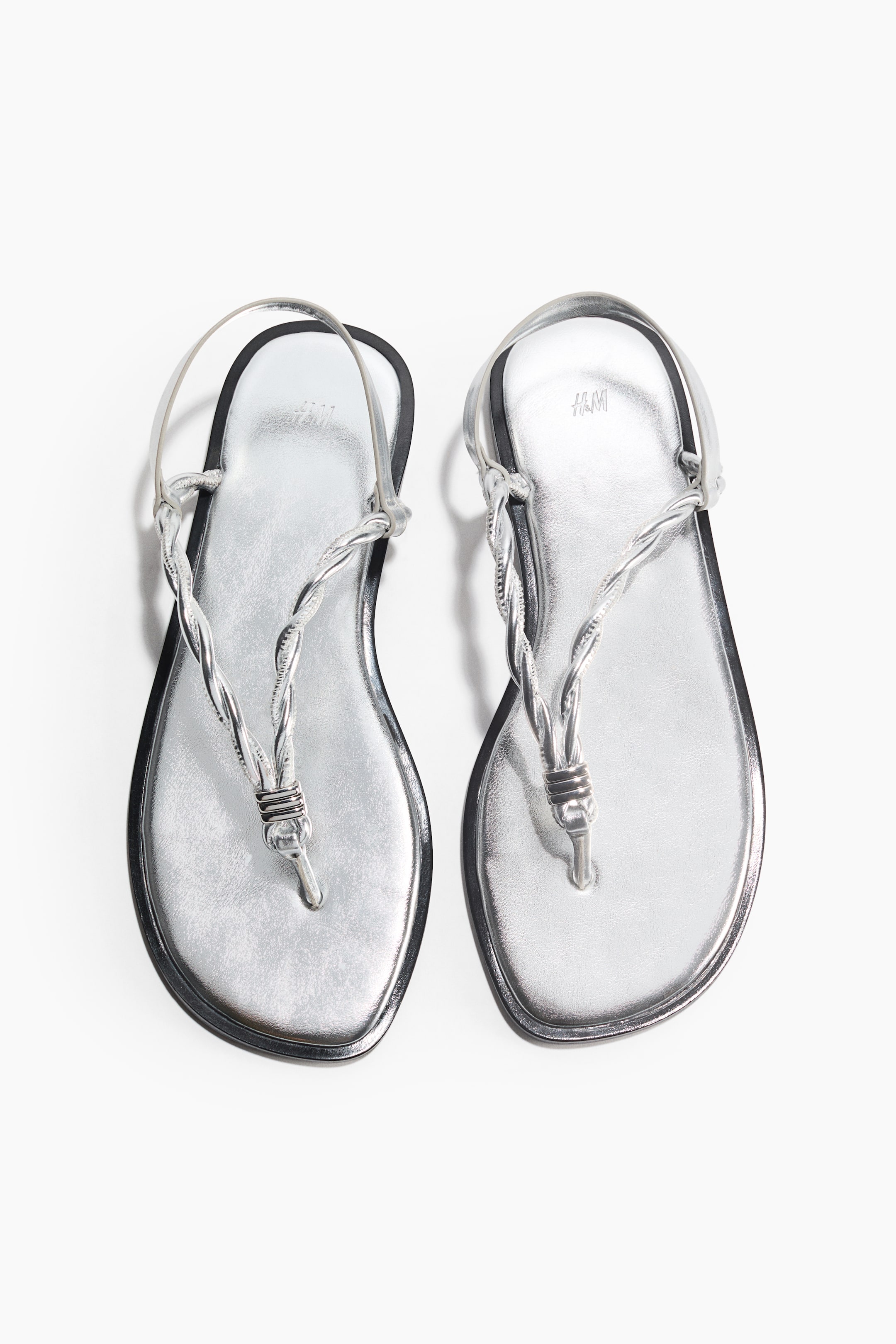 View larger image: Two silver thong sandals are showcased with metallic silver footbeds and black-edged soles. Each sandal features a twisted silver thong strap extending to a toe post and a thin silver ankle strap.