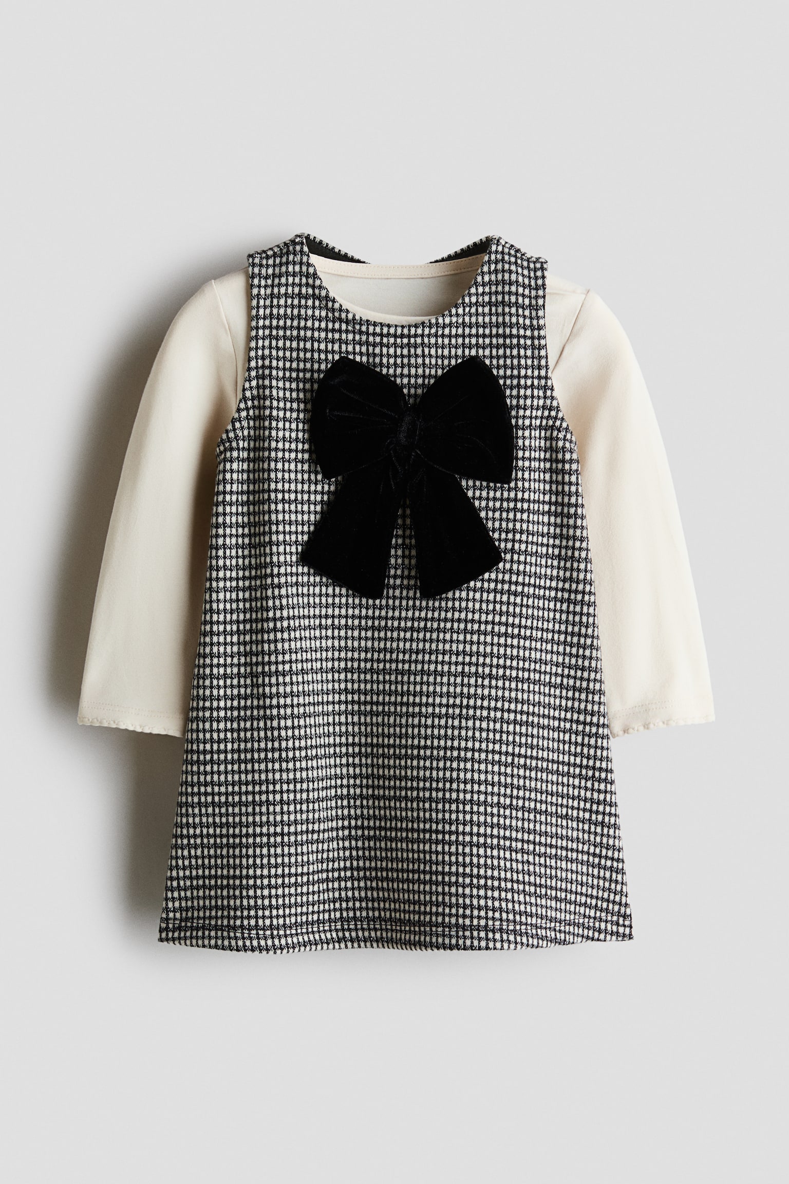 A childâs two-piece dress set is shown from the front, featuring a sleeveless black-and-white houndstooth pinafore dress layered over a cream-coloured long-sleeve top. The dress has an A-line silhouette with a large black velvet bow centered on the chest.
