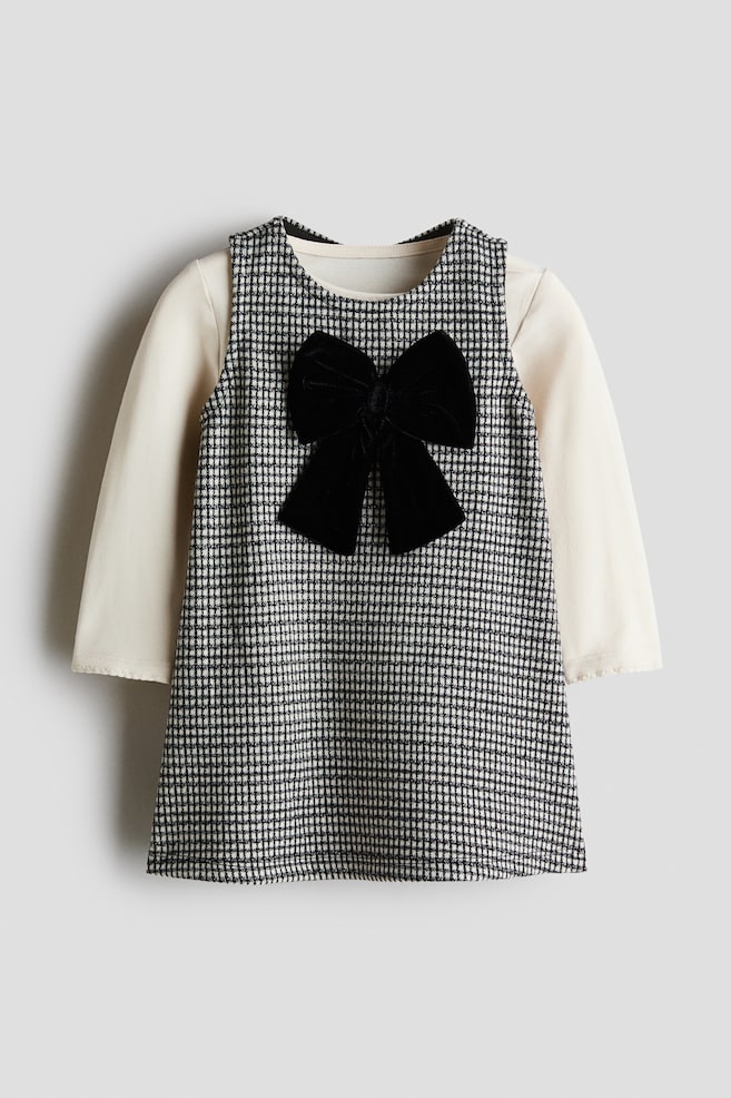 Een tweedelige jurkenset voor een kind is van voren afgebeeld en laat een mouwloze zwart-witte houndstooth pinafore-jurk zien, gecombineerd met een crÃ¨mekleurige top met lange mouwen. De jurk heeft een A-lijnsilhouet met in het midden van de borst een grote zwarte fluwelen strik. - 1