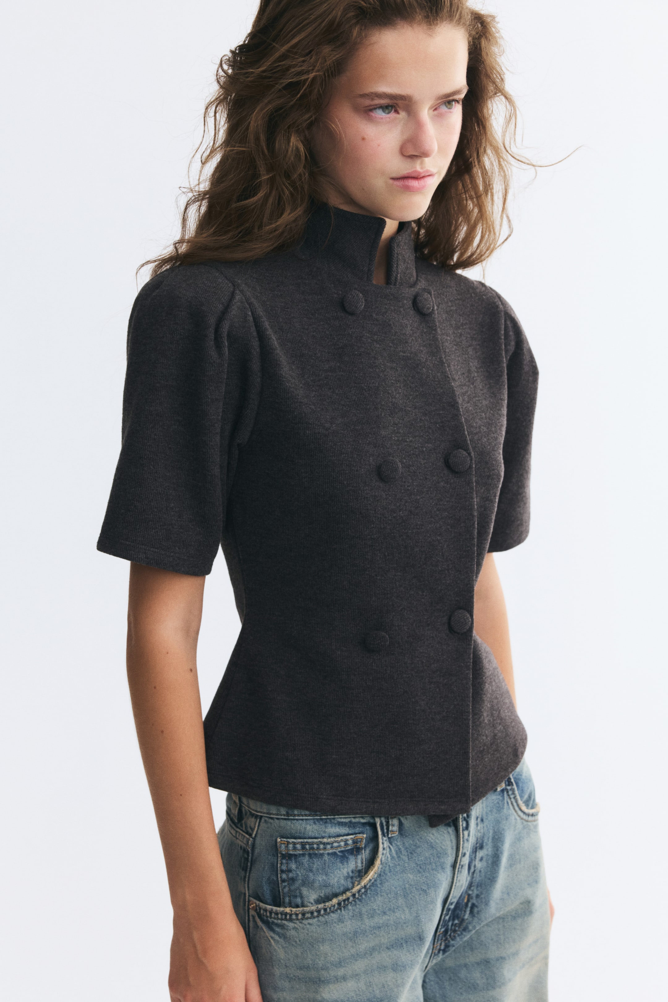 Größeres Bild ansehen: Ein dunkelgraues, figurbetontes Oberteil aus feinem Rippjersey wird von dem Model getragen und verfügt über einen asymmetrischen Stehkragen, kurze Puffärmel und eine zweireihige Vorderseite mit verdeckten Knöpfen. Das Oberteil ist mit hellblauen Denim-Jeans kombiniert.