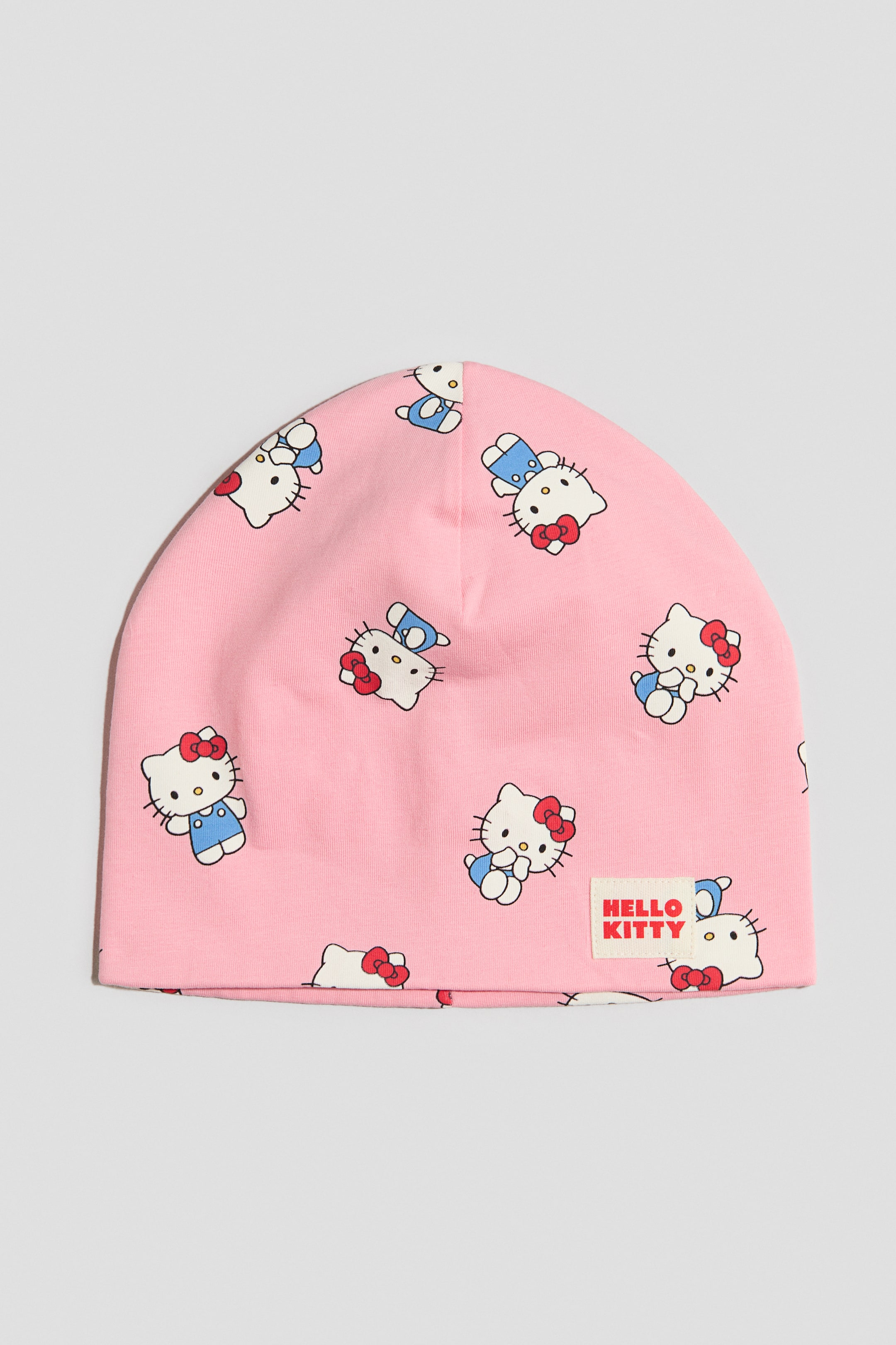 Zobacz większy obraz: Różowa czapka beanie leży na płasko, prezentując powtarzający się wzór z postaciami Hello Kitty, z których niektóre mają niebieskie ogrodniczki, a inne czerwone kokardki. Na froncie znajduje się prostokątna tkana metka z napisem „HELLO KITTY” czerwonym tekstem.