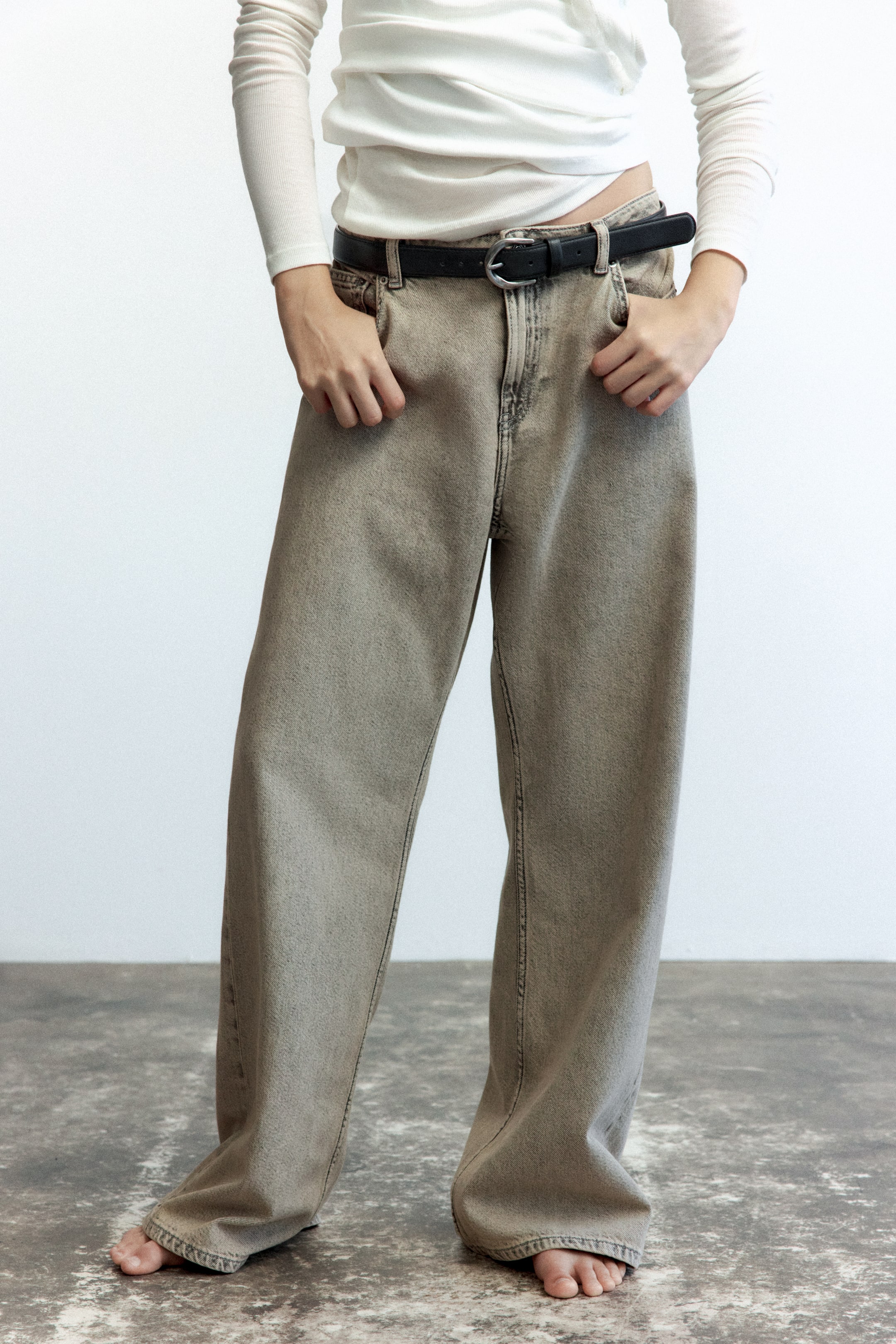 View larger image: Loose-fitting, beige-washed denim jeans with a wide-leg silhouette are worn by the model, who has their hands in the front pockets. The jeans are styled with a black leather belt and a long-sleeved white top.