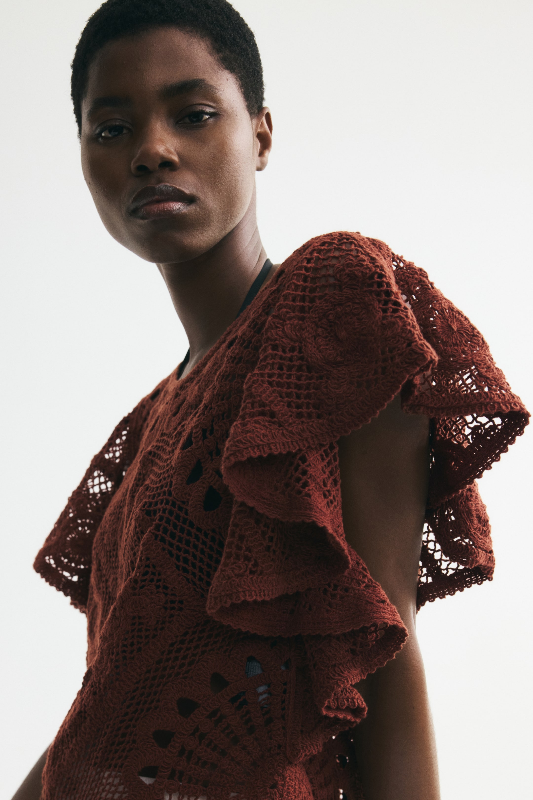 Größeres Bild ansehen: Ein Oberteil im Häkel-Look-Strick mit Rundhalsausschnitt und voluminösen Flatterärmeln wird von dem Model getragen. Ein dunkler Unterwäscheträger ist durch das offene Strickmuster des Oberteils sichtbar.