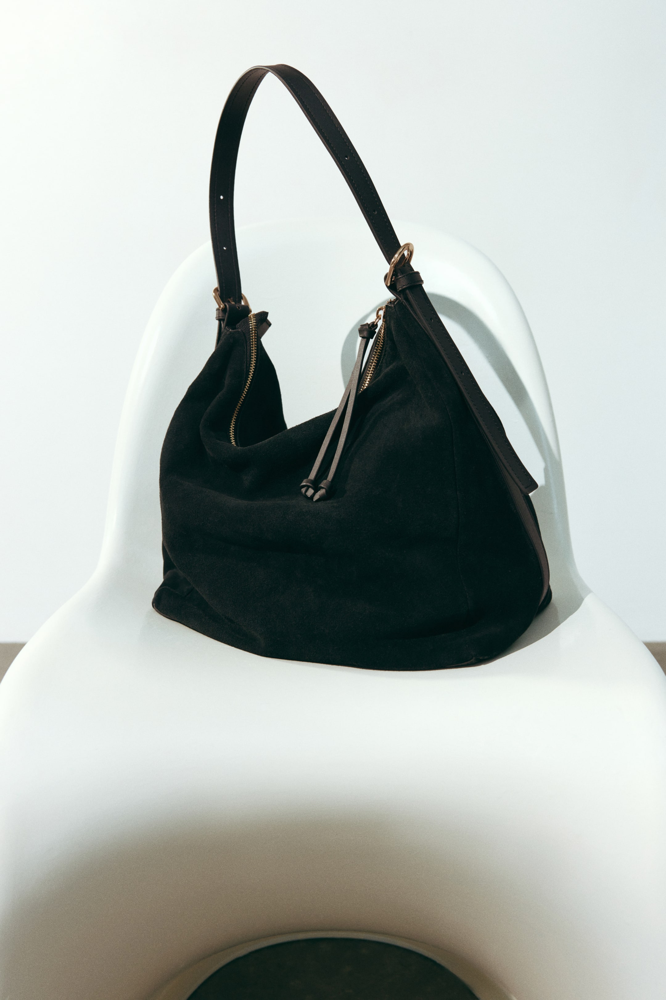 Größeres Bild ansehen: Eine schwarze Wildleder-Umhängetasche liegt auf einem weißen Stuhl und zeigt ihr lässig fallendes Design, einen goldfarbenen Reißverschluss mit langem, geknotetem Zipperband sowie einen verstellbaren Schulterriemen aus Leder mit Metallschnallen.