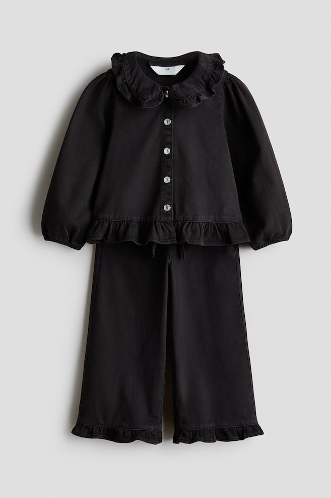 A two-piece black set is facing the front, featuring a long-sleeved button-up shirt and matching wide-leg trousers. The shirt is displaying a ruffled Peter Pan collar, silver button closures, and a ruffled hemline, while the trousers are showing matching ruffled cuffs at the bottom of each leg. - 1