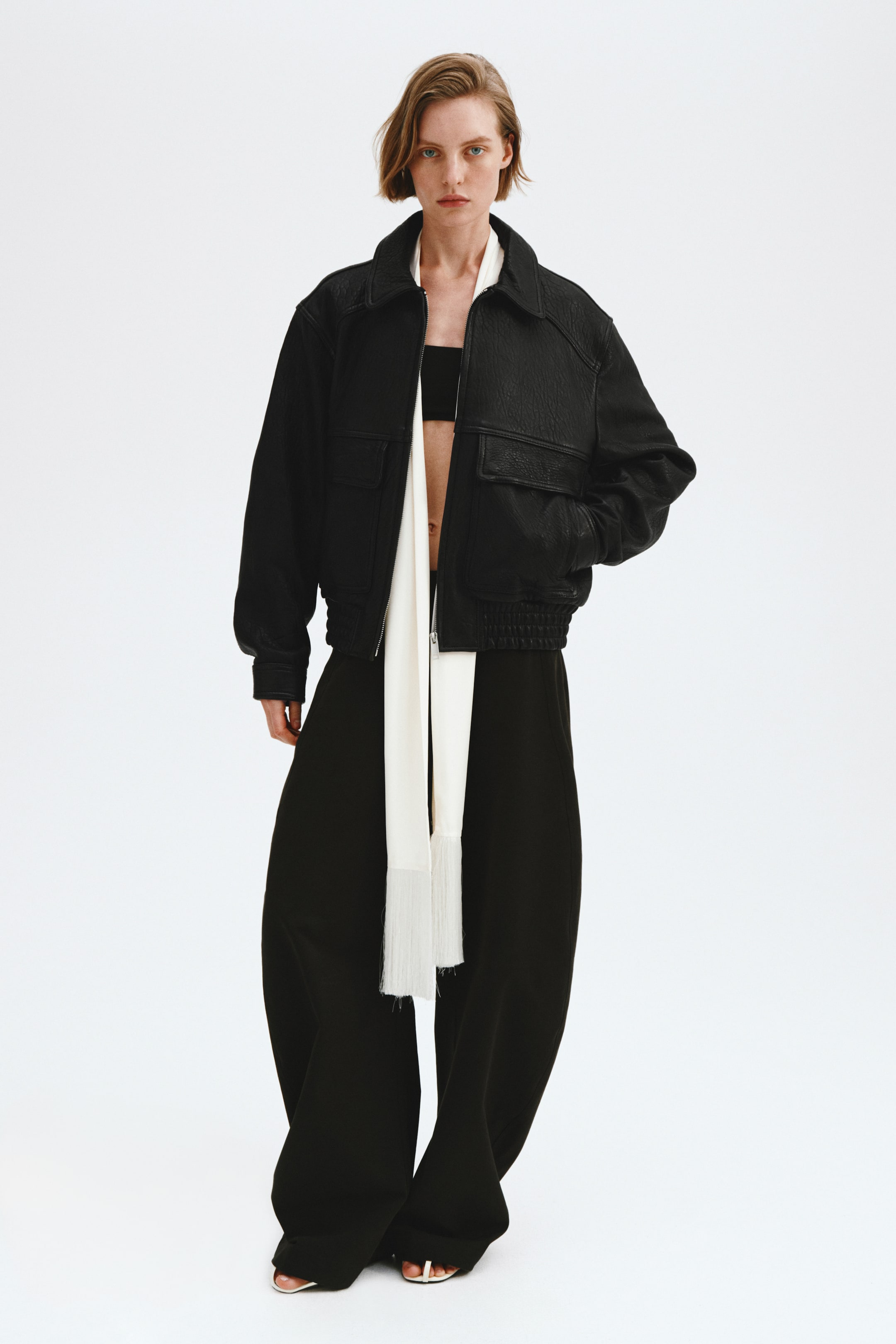 Größeres Bild anzeigen: Eine schwarze, strukturierte Bomberjacke wird von vorne präsentiert, mit einem spitzen Kragen, zwei Brusttaschen mit Klappen und elastischen Bündchen und Saum. Sie wird offen über einem schwarzen Bandeau-Top getragen, mit einem langen weißen Fransenschal, der darunter drapiert ist und über weit geschnittene schwarze Hosen fällt.