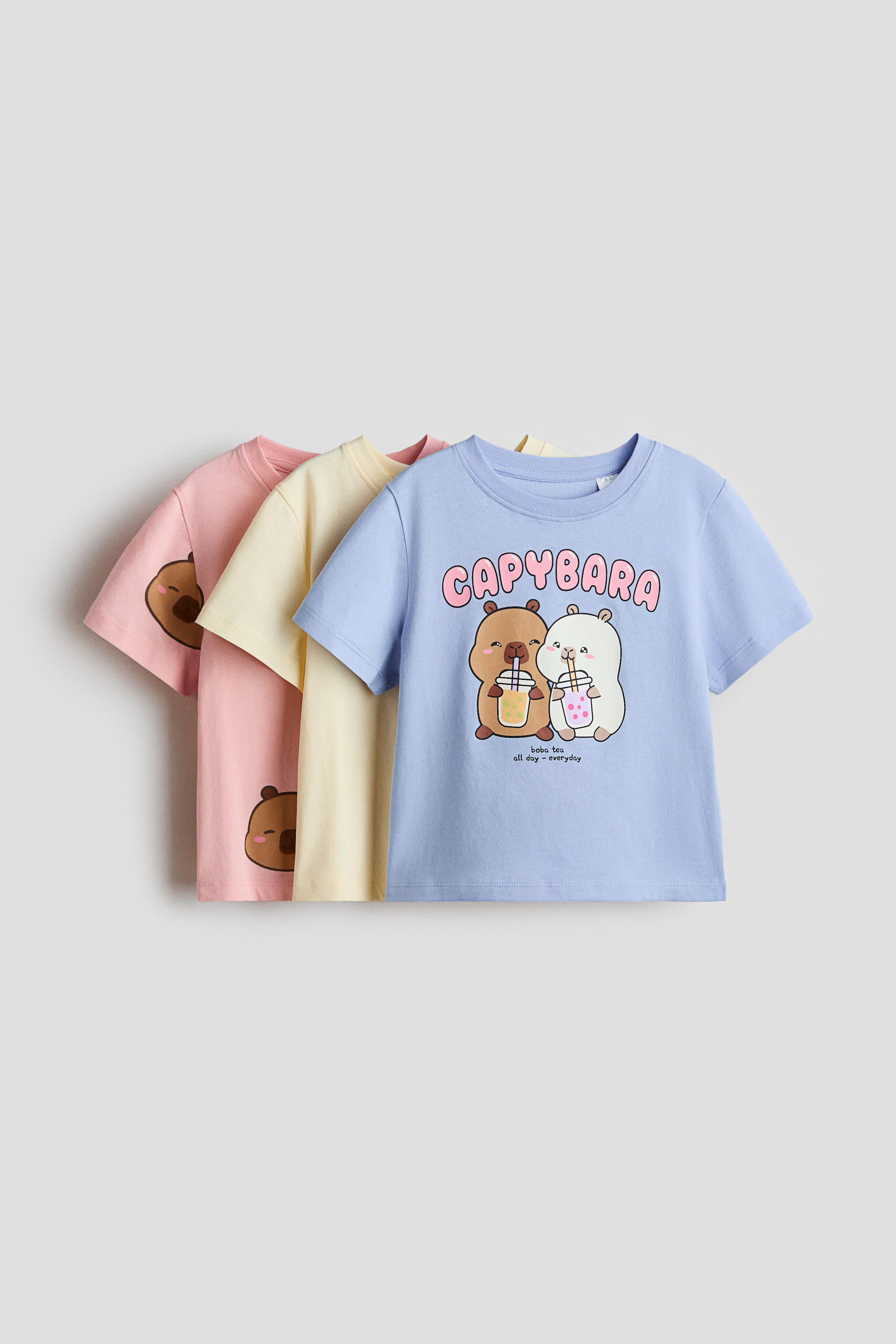 View larger image: Three short-sleeved jersey tops with crew necks are layered, with the front light blue top displaying a graphic print of two cartoon capybaras holding bubble tea, "CAPYBARA" text, and "boba tea all day everyday" text. The light yellow and light pink tops are partially visible behind, each featuring a brown cartoon capybara head print on the lower front.