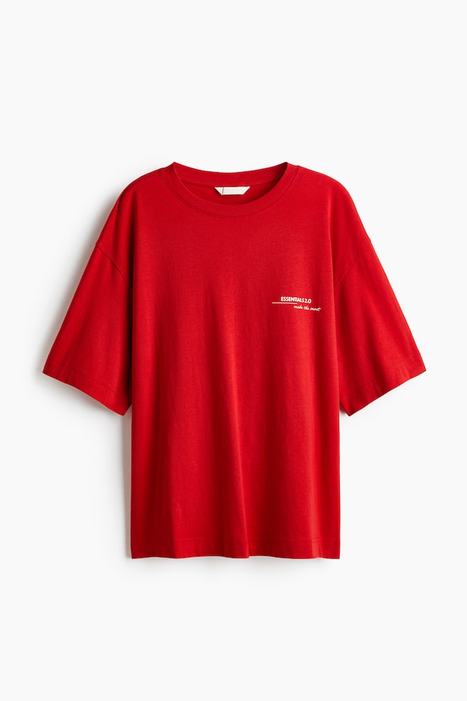 A front-facing red t-shirt is featuring a crew neck and short, loose-fitting sleeves with dropped shoulders. White text "ESSENTIALS 2.0 make the most" is appearing on the left chest. - 1