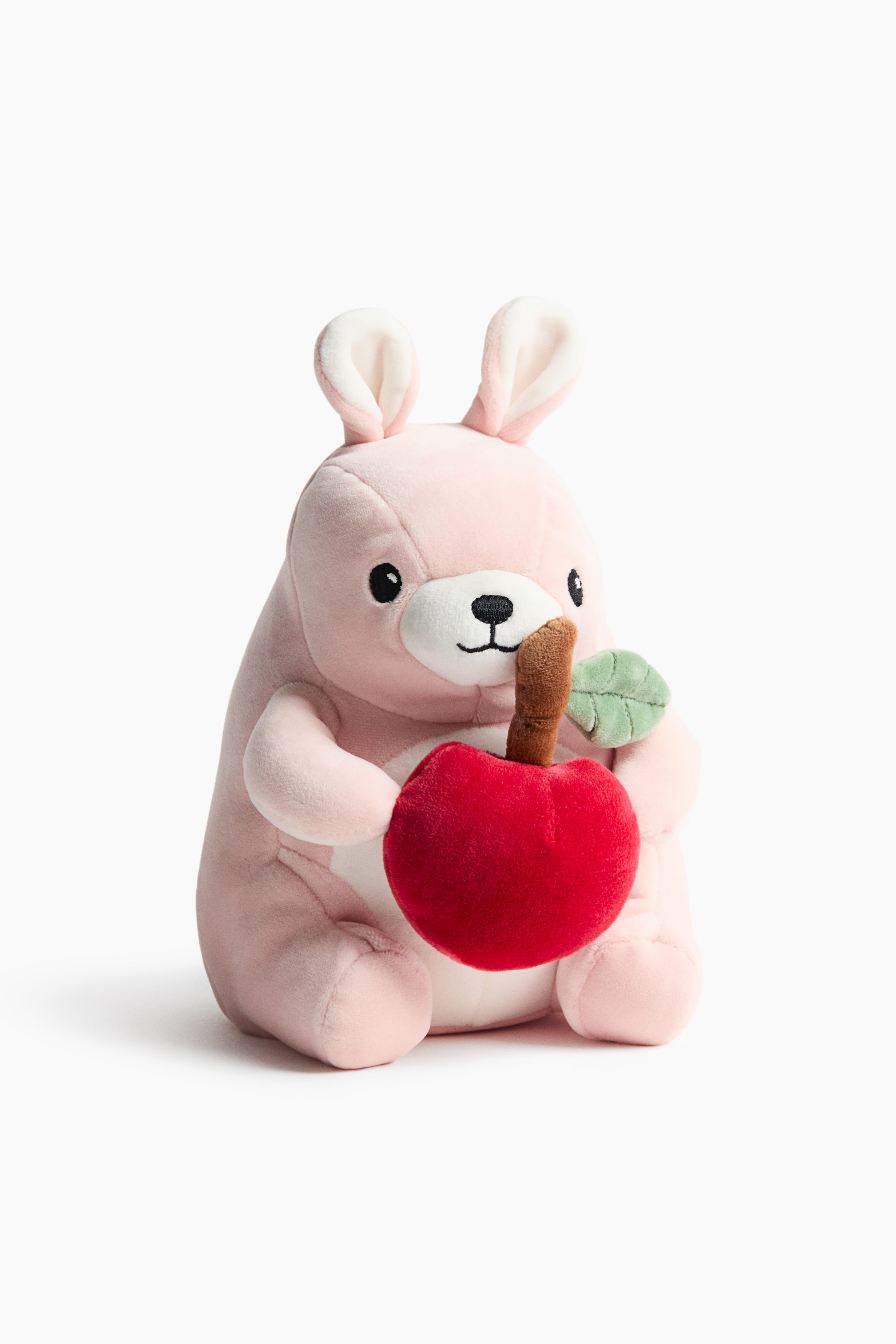 Ver imagen más grande: Un peluche suave rosa está sentado y de frente mientras sostiene una gran manzana de peluche roja con un tallo marrón y una hoja verde. El juguete presenta ojos negros bordados, un hocico blanco y un parche blanco en el vientre, así como orejas erguidas rosas y blancas.