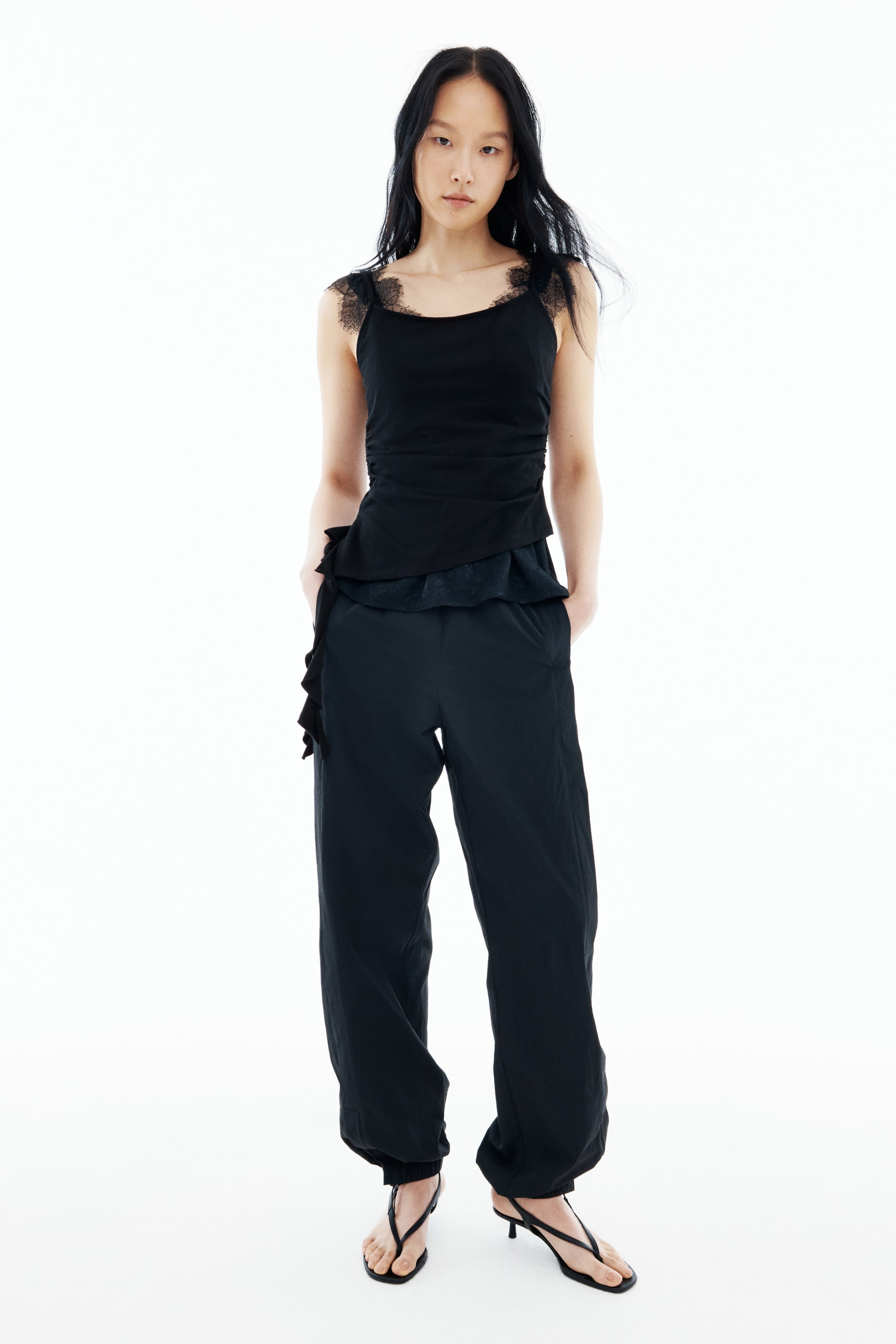 View larger image: A black camisole top with lace-trimmed straps and an asymmetrical, ruffled hem is being worn with black relaxed-fit pants featuring gathered ankles. The model is standing with hands in the pants pockets, while black thong-style sandals are being worn.