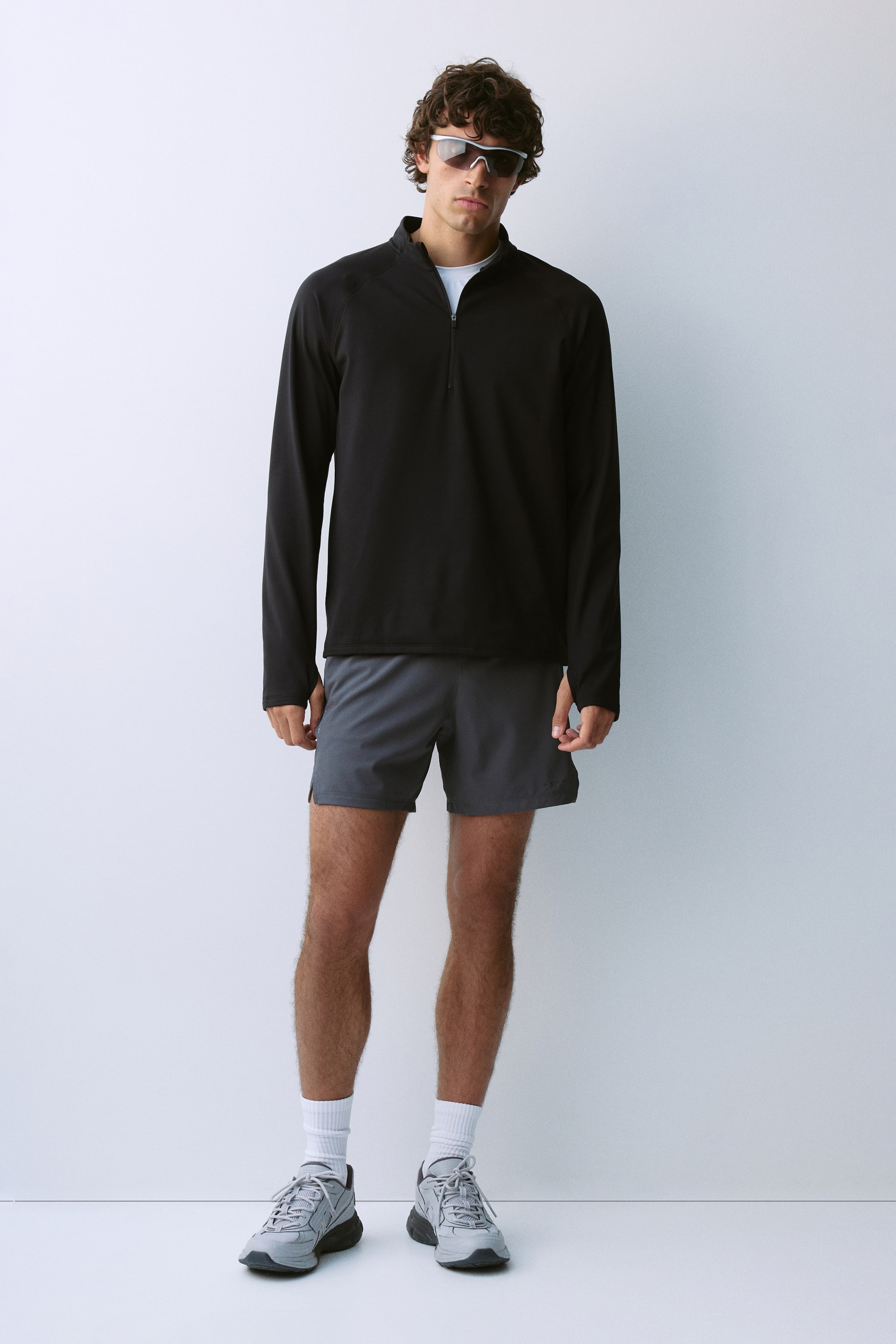 View larger image: A black, regular-fit half-zip activewear top is worn by the model. The long-sleeved top features a high collar with a partially open zipper revealing a white T-shirt collar underneath, and is styled with grey athletic shorts.