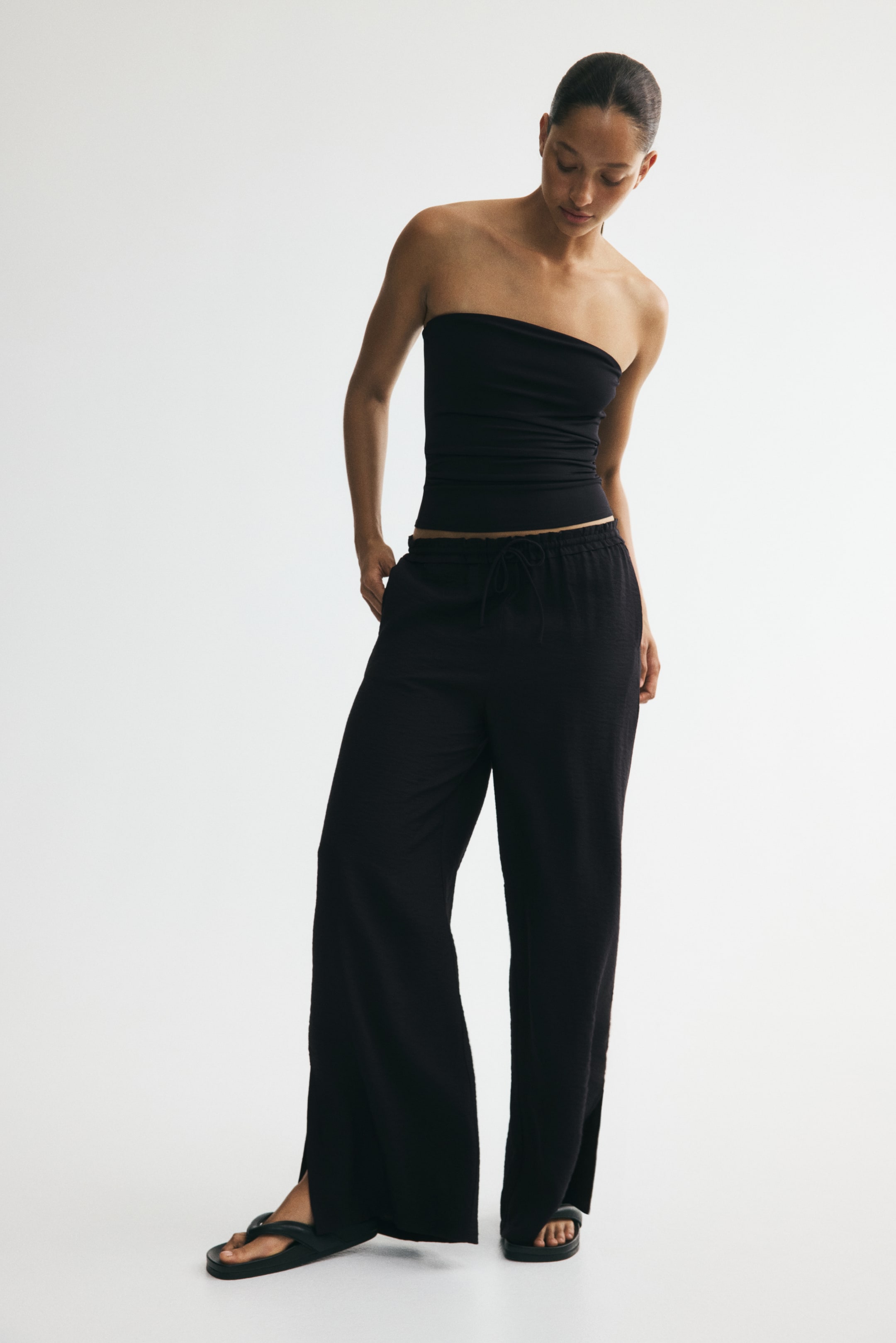 Größeres Bild ansehen: Schwarze, locker sitzende Hose aus einem sanft geknitterten Viskose-Mischgewebe mit leichtem Glanz, ausgestattet mit einem elastischen, mit Rüschen besetzten Bund und einer schmalen Kordel sowie weiten Beinen mit Schlitzen an den Säumen. Das Model trägt die Hose und steckt die Hände in die Taschen.