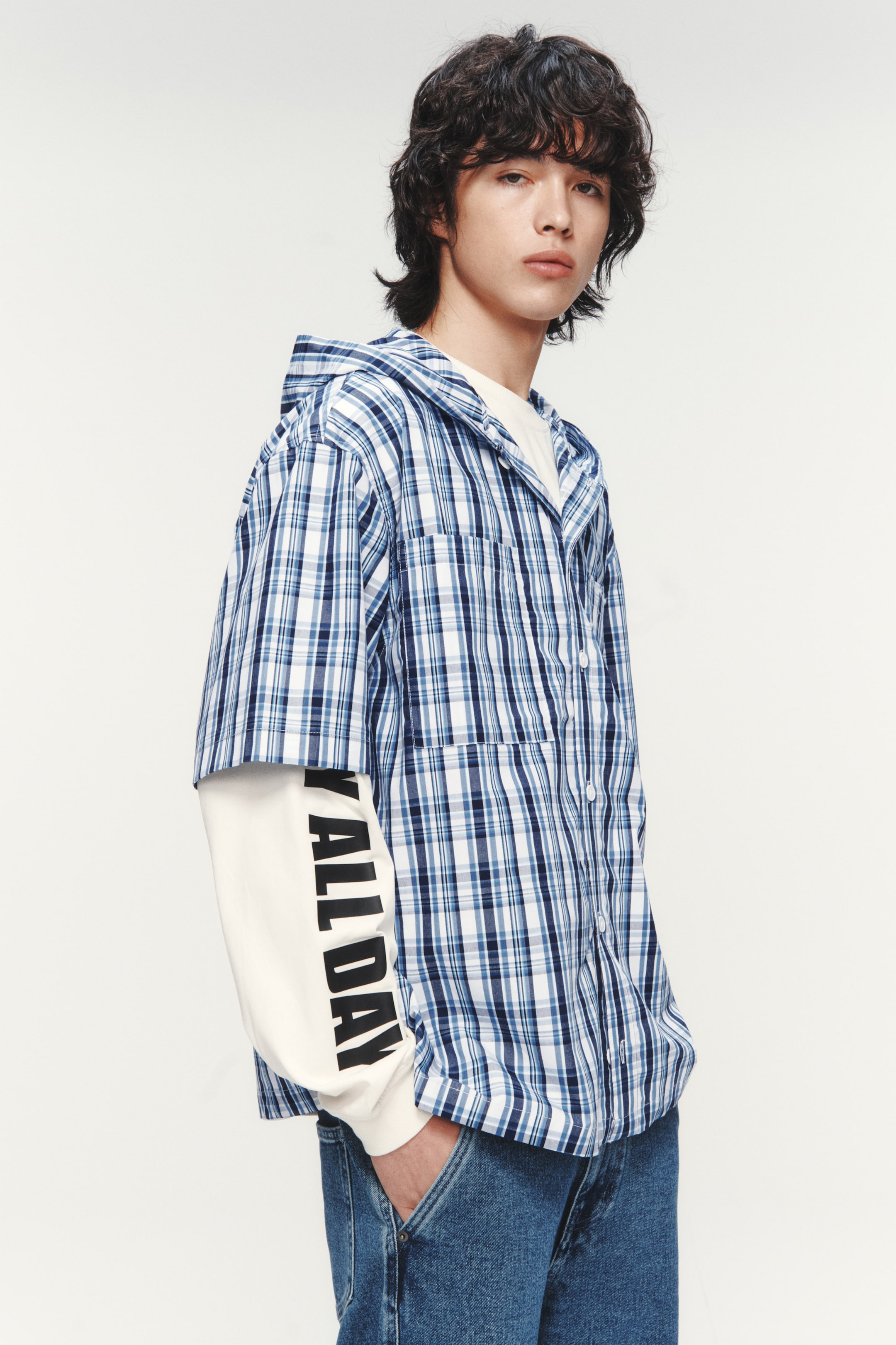 Xem ảnh lớn hơn: Áo sơ mi có mũ trùm đầu ngắn tay kẻ sọc xanh trắng, với thiết kế cài nút phía trước và túi ngực, hơi nghiêng về bên phải. Áo được phối cùng áo dài tay màu kem, có chữ 'ALL DAY' in dọc màu đen trên tay áo bên trái.