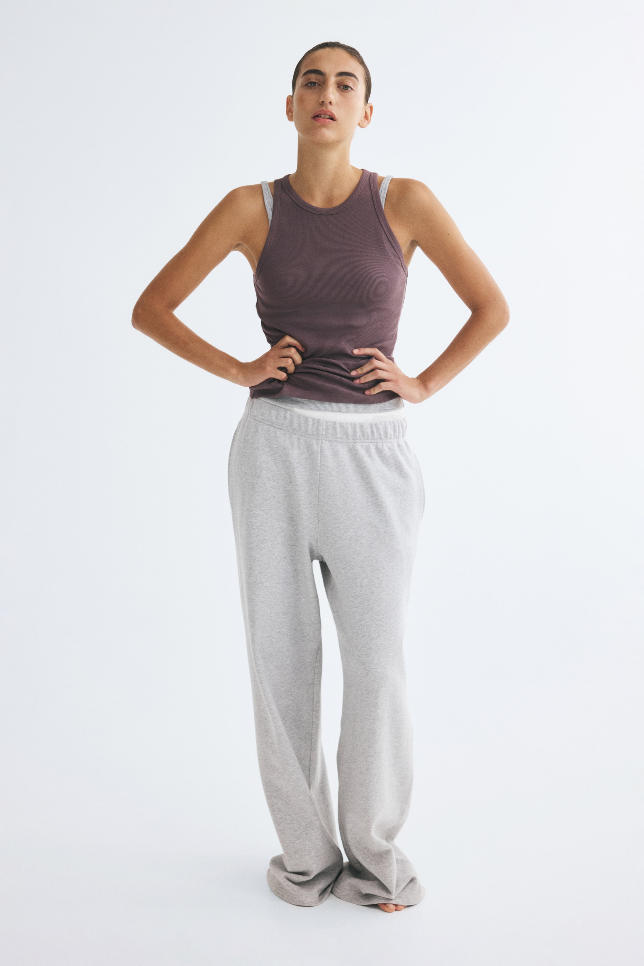 View larger image: A ribbed cotton jersey tank top in a dark purple shade, featuring a round neckline and a straight hem, is worn by the model. The tank top is layered over a light grey tank top and styled with loose grey sweatpants.