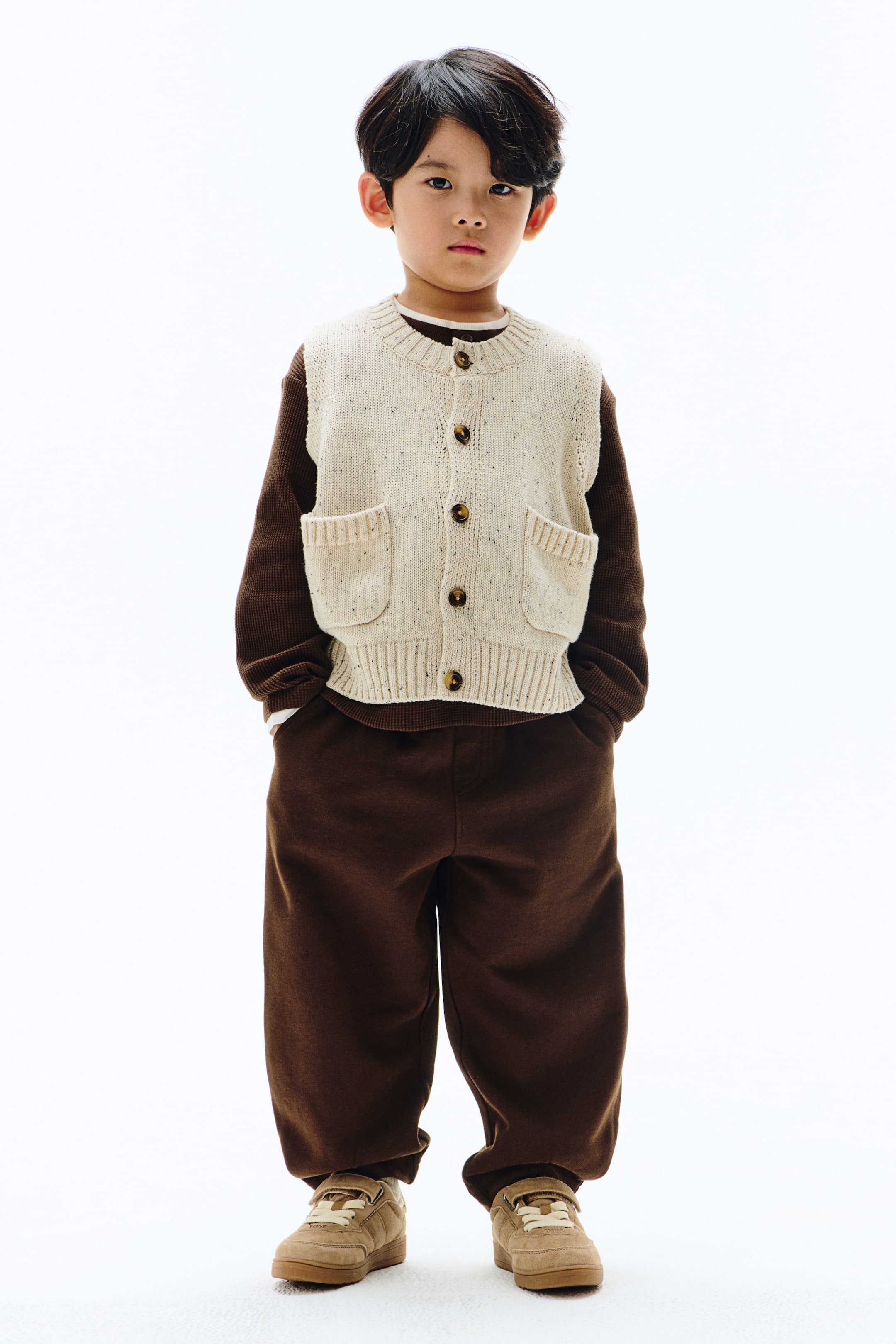이미지 확대 보기: 어린이의 옷차림은 크림색 반점 니트 조끼와 버튼 앞부분, 두 개의 패치 포켓, 그리고 칼라, 팔 구멍, 밑단에 리브 디테일이 특징입니다. 이 조끼는 짙은 갈색의 긴 소매 와플 니트 셔츠 위에 레이어드되어 있으며, 짙은 갈색 트레이닝 바지와 베이지색 스니커즈로 완성된 코디입니다.