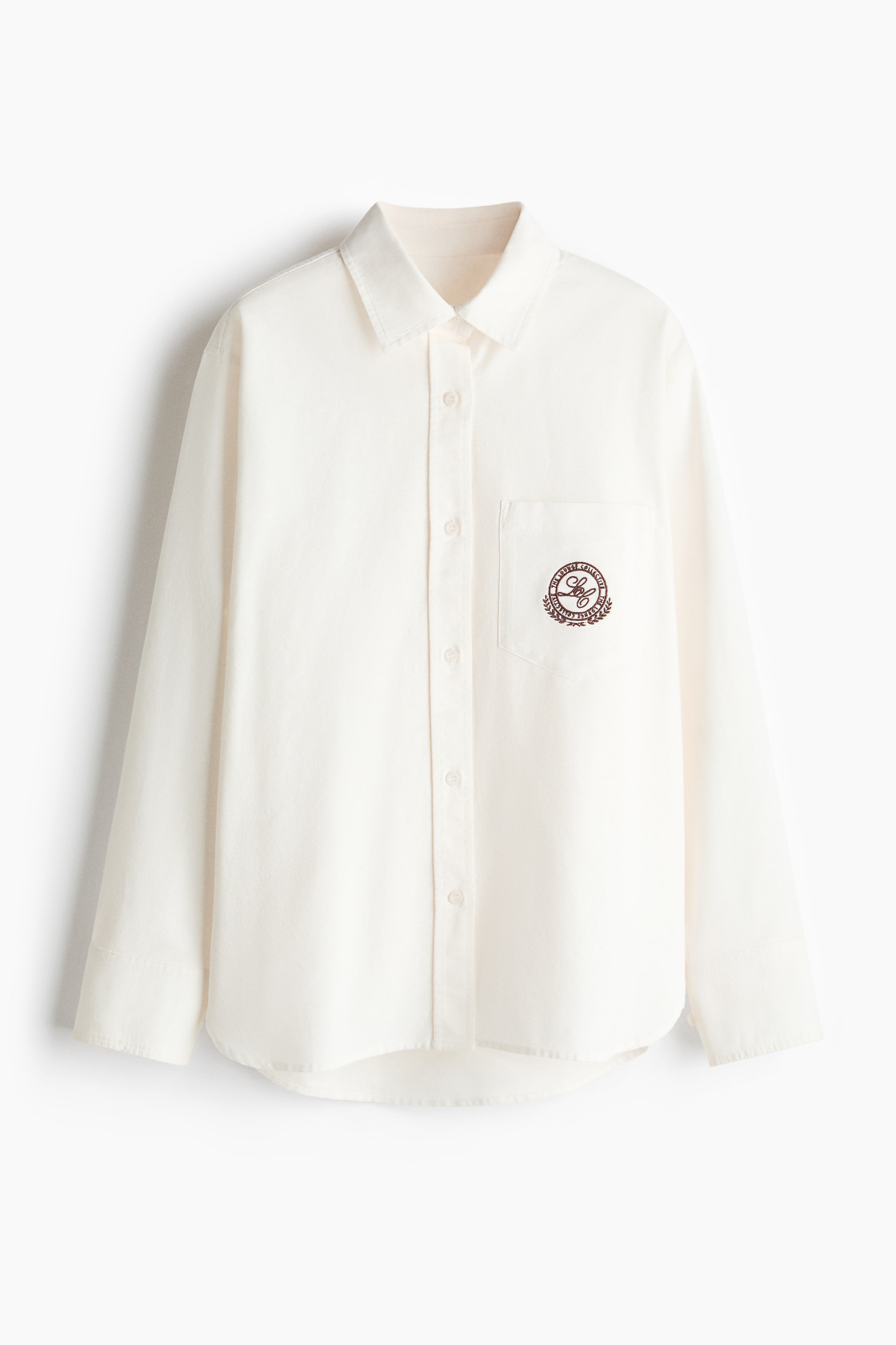 View larger image: A cream-coloured shirt is showcasing its front, boasting a pointed collar, a full-button placket, and a patch pocket on the left chest embellished with a dark brown circular embroidered emblem.