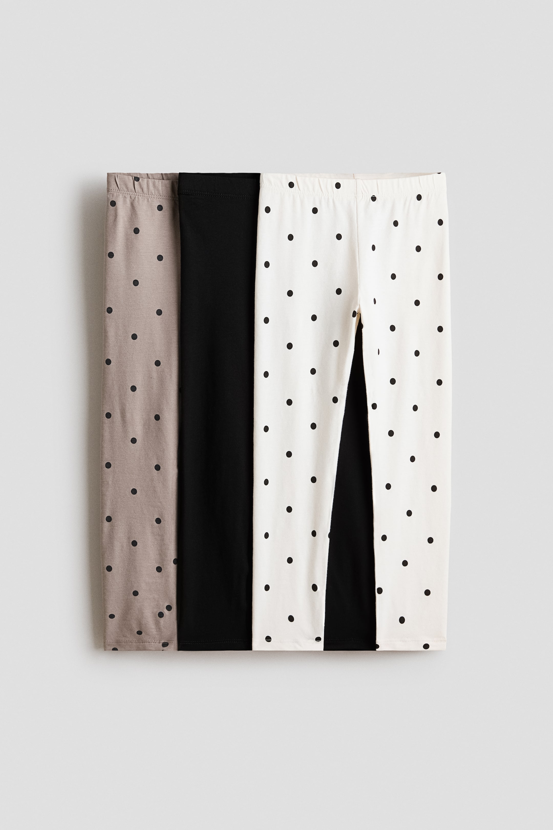 View larger image: Three pairs of leggings are showcased, arranged flat in a vertical layout. The left pair features a taupe hue with black polka dots, the middle pair is a classic solid black, and the right pair is cream adorned with black polka dots. Elasticated waistbands are evident on both the taupe and cream pairs.