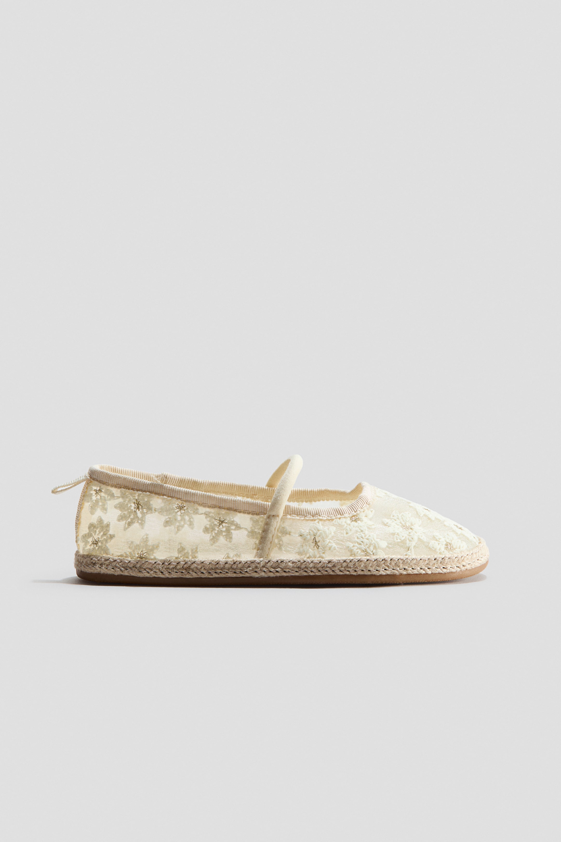 View larger image: A cream espadrille shoe is showcased in a side view, featuring a rounded toe and an upper crafted from sheer fabric adorned with embroidered floral patterns. A slim, elastic strap stretches across the foot, a small loop extends from the back heel, and braided jute trim is visible around the soles.