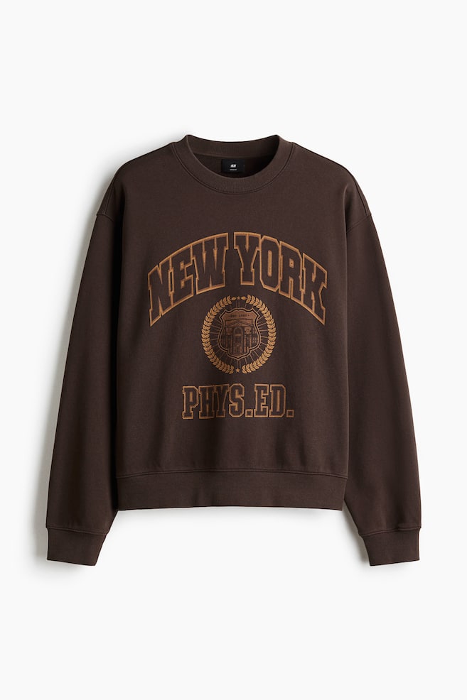 A brown sweatshirt is shown from the FRONT, featuring a loose fit, a round, rib-trimmed neckline, dropped shoulders, long sleeves with wide ribbed cuffs, and a wide ribbed hem. A lighter brown graphic print is visible across the chest, displaying "NEW YORK" in an arch, a crest, and "PHYS.ED." - 1